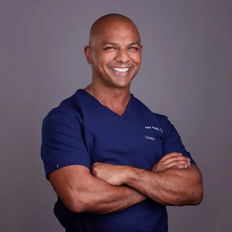 A man in a navy medical scrub top, arms crossed, smiling at the camera against a plain gray background.