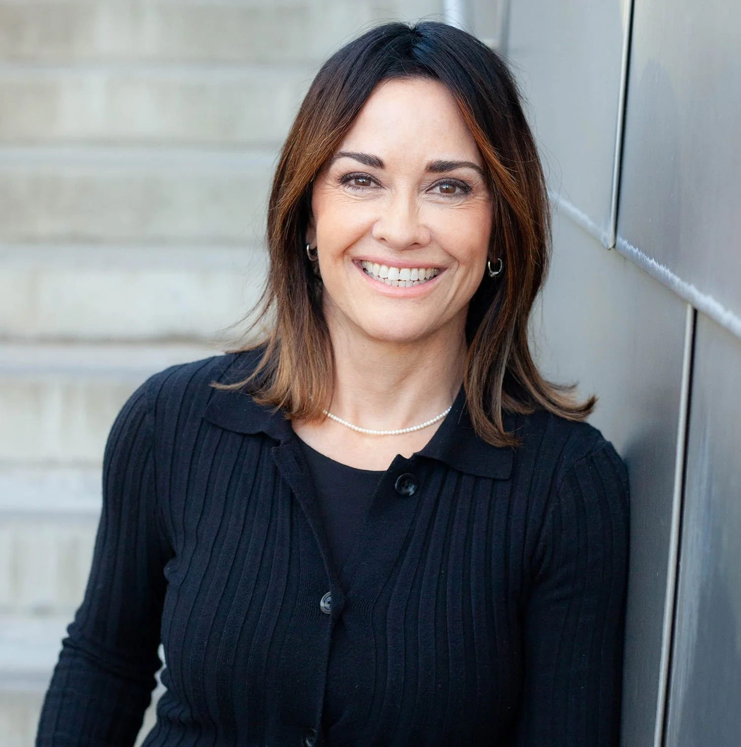 Portrait of a smiling woman with shoulder-length brown hair, wearing a black button-up shirt, standing outdoors against a concrete staircase and a metallic wall.