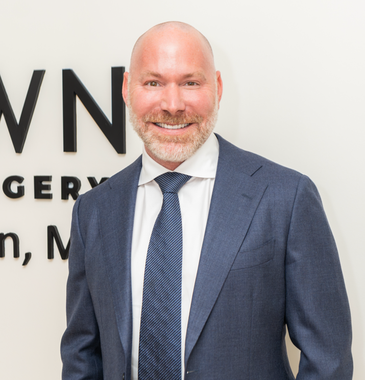 A smiling man in a blue suit with a white shirt and blue tie, standing in front of a white wall with partial black text.