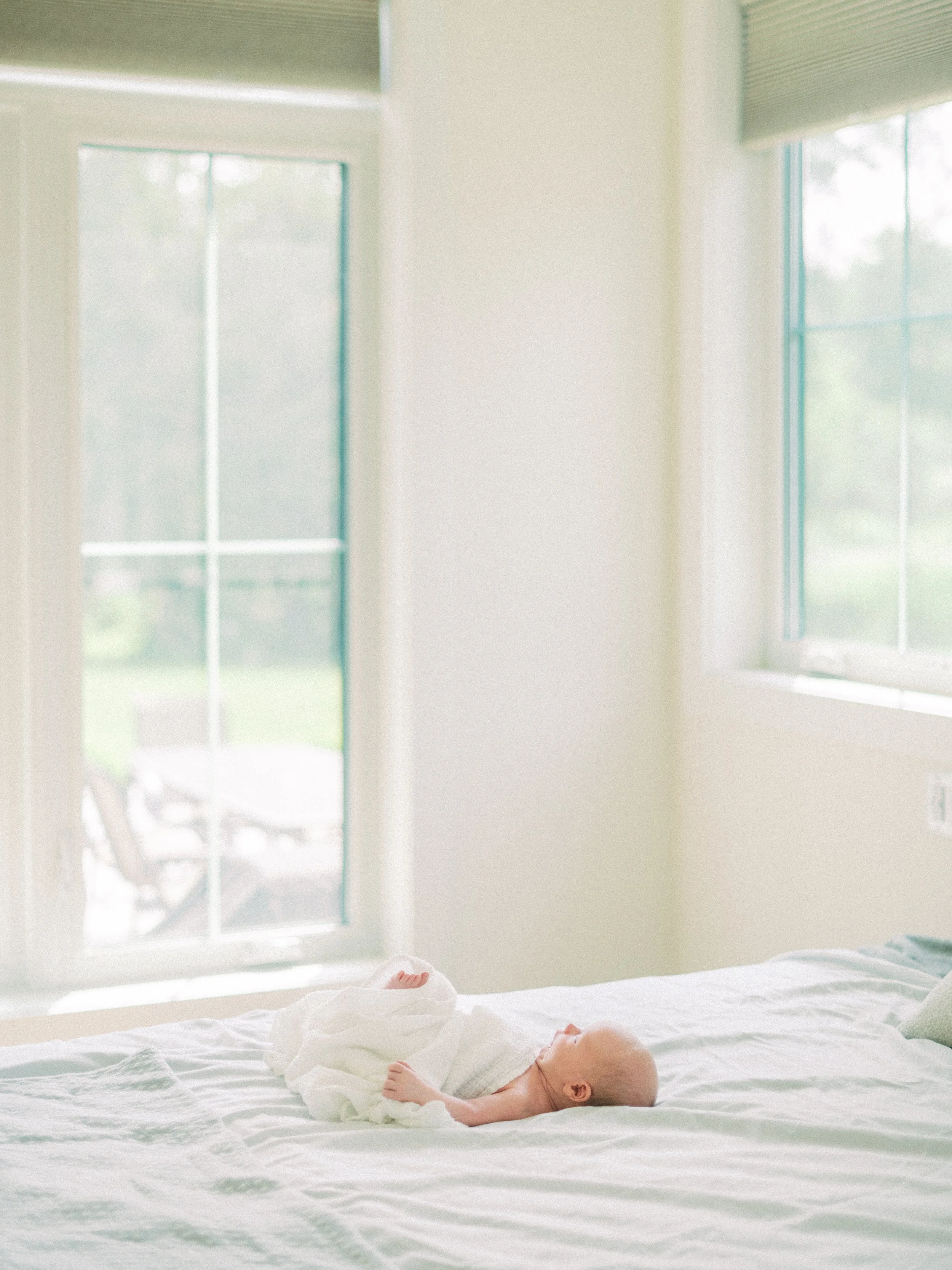 A sleeping baby lying on a bed in a bright bedroom with large windows and white walls.
