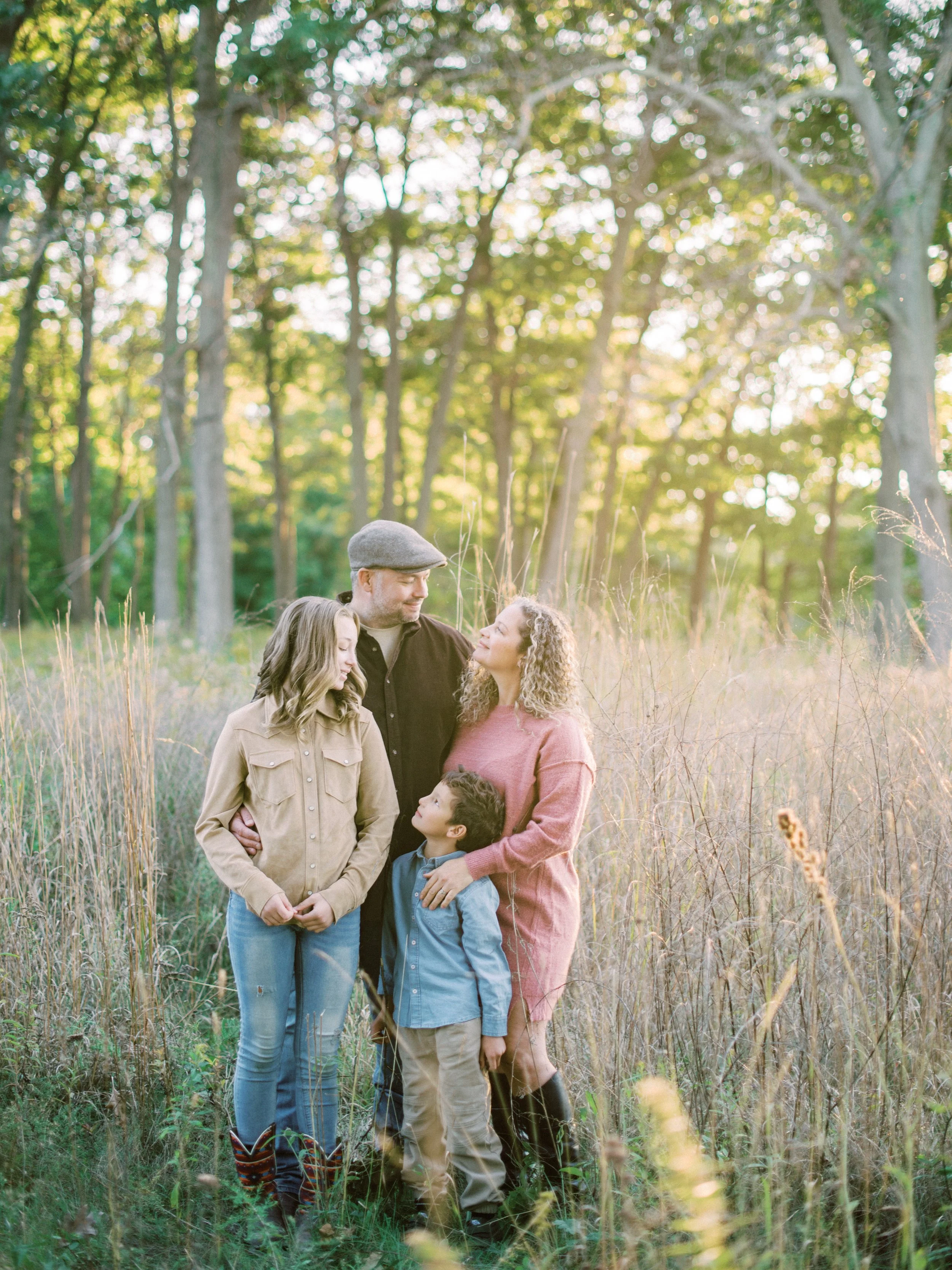 A family of four, including a mother, father, daughter, and son, standing in a forest clearing during sunset, embracing and looking at each other.
