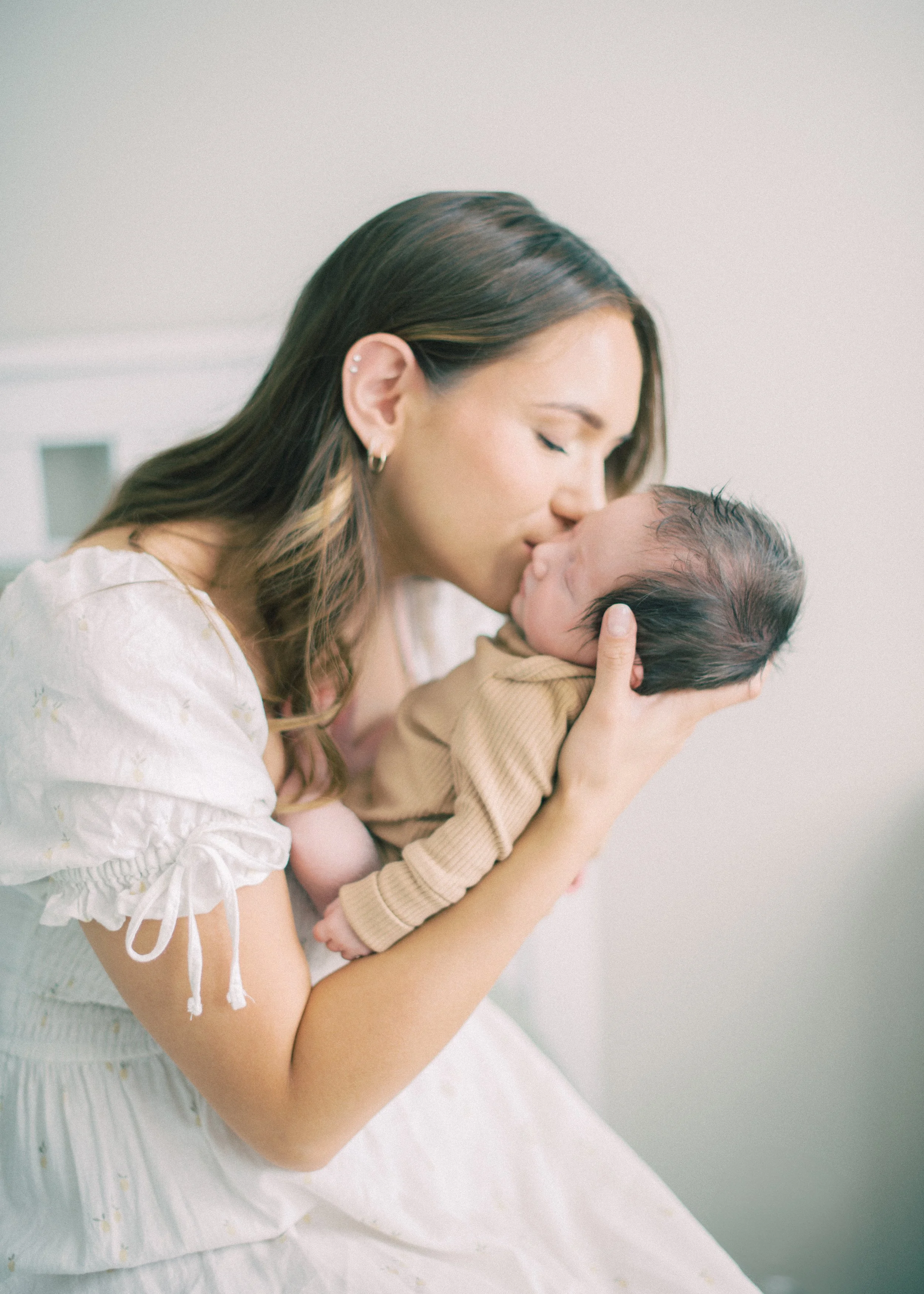 A woman with brown hair and piercings gently holding and kissing a newborn baby with dark hair, indoors with a plain, light-colored background.