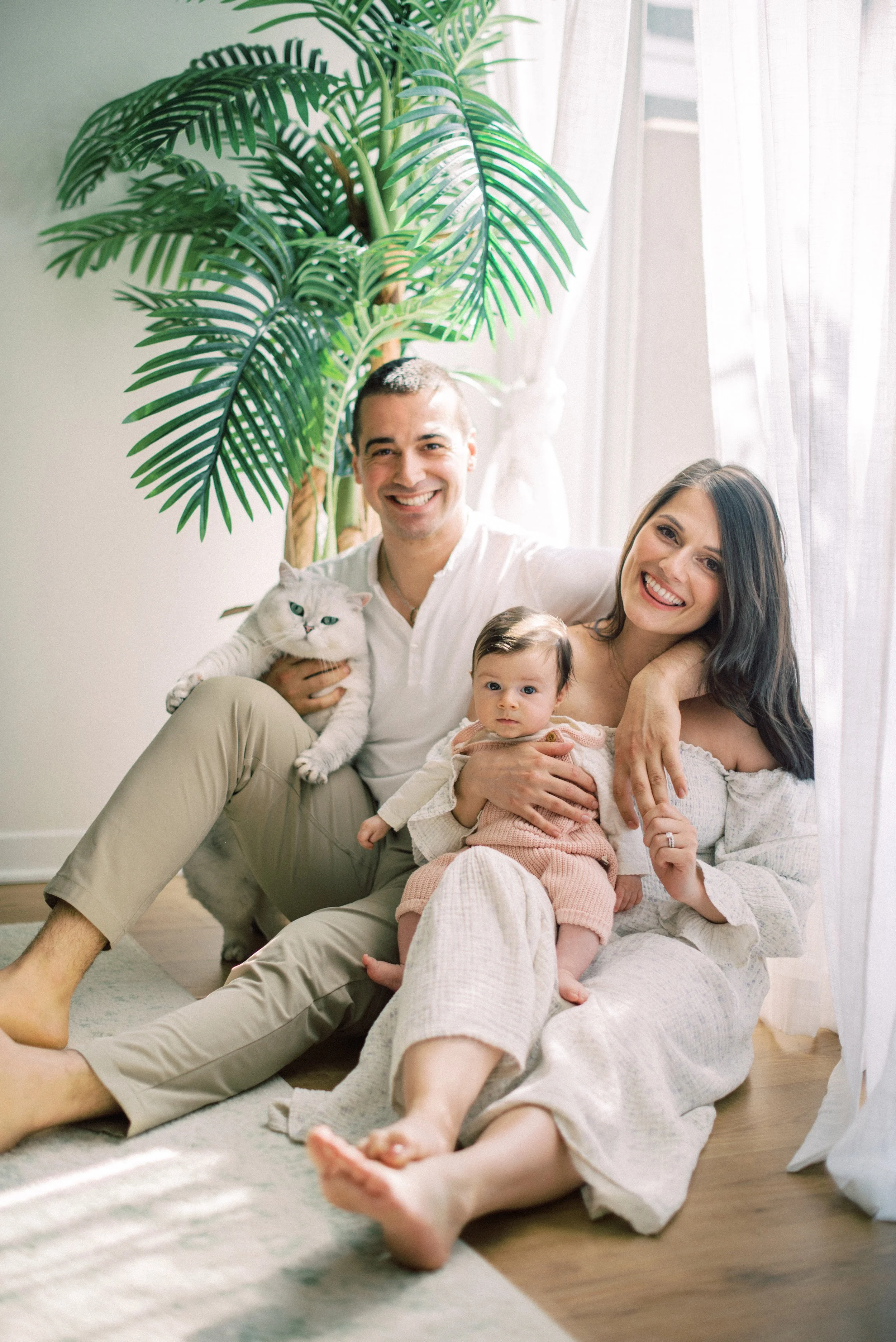 A happy family of four, including a woman, man, baby, and a cat, sitting on the floor near windows with curtains in a bright room, with a large potted plant in the background.