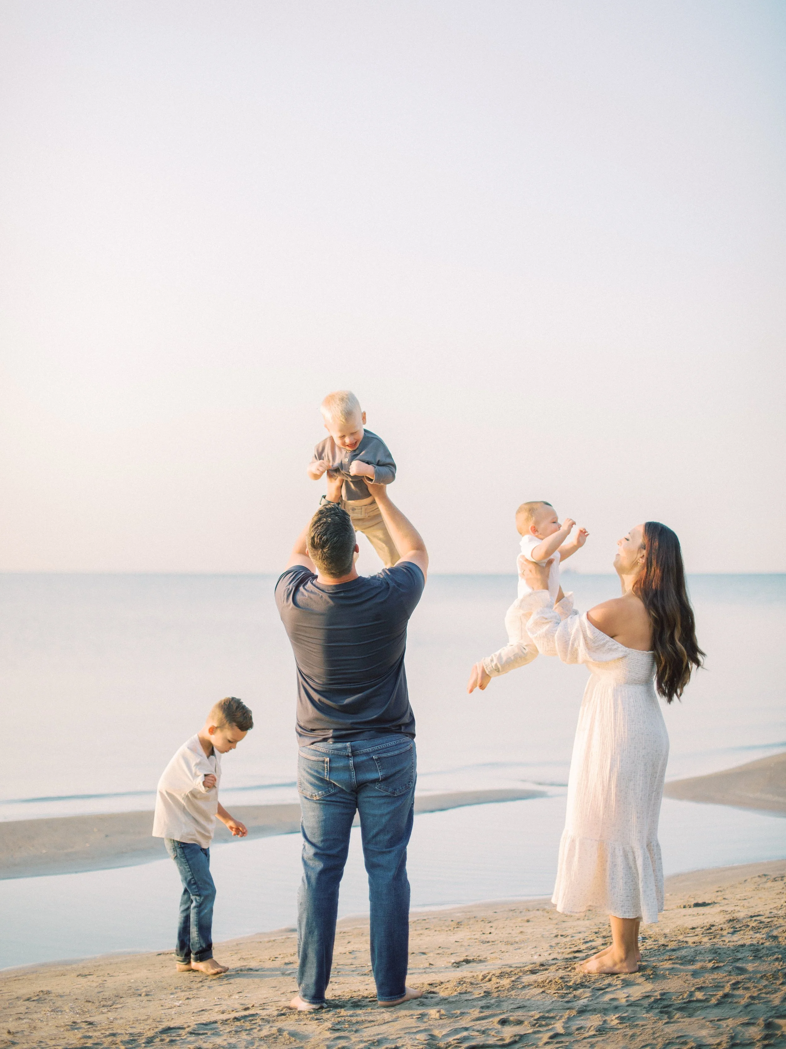 A family enjoying a day at the beach. A man is lifting a young child above his head, while a woman is holding a smiling baby. Another boy is looking down at the sand.
