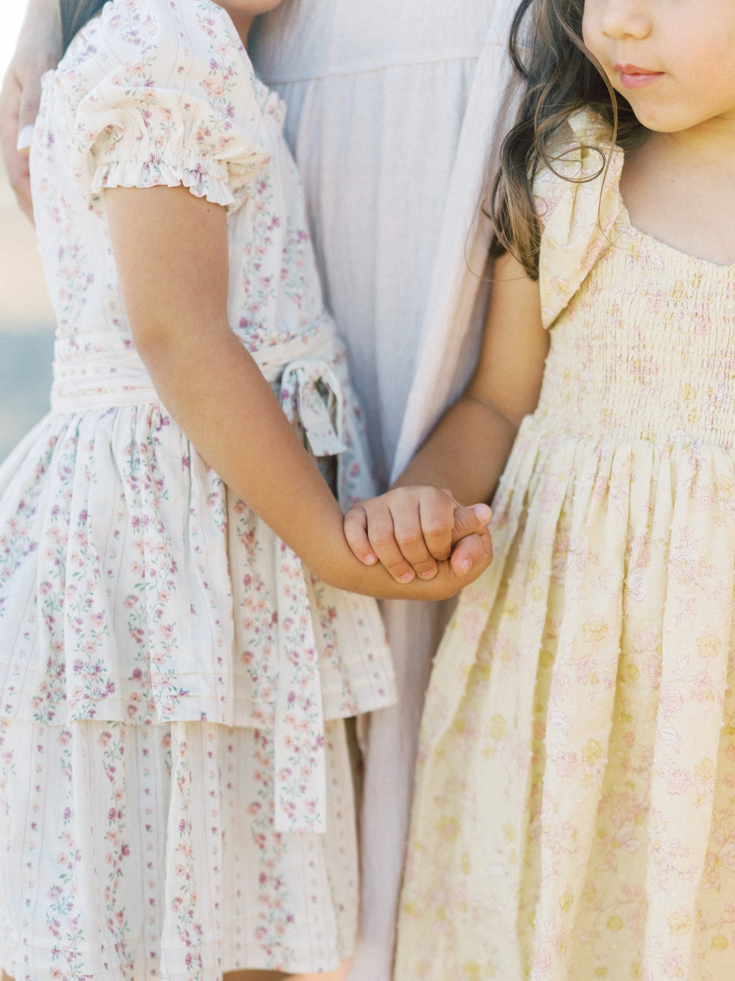 Two young girls holding hands, wearing light-colored floral summer dresses, standing outdoors.