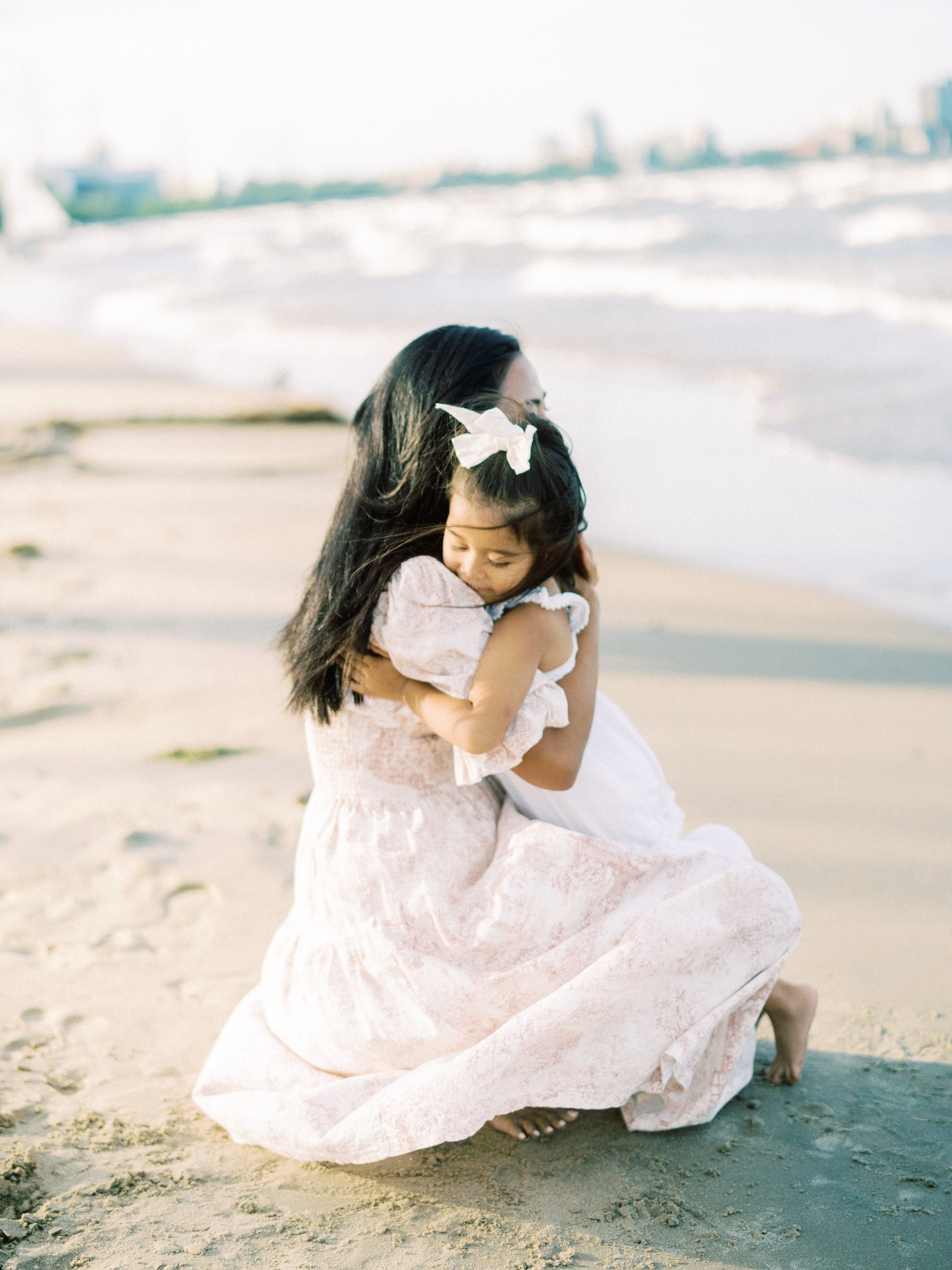 A woman and a young girl hugging on the beach near the ocean, with city buildings in the background.