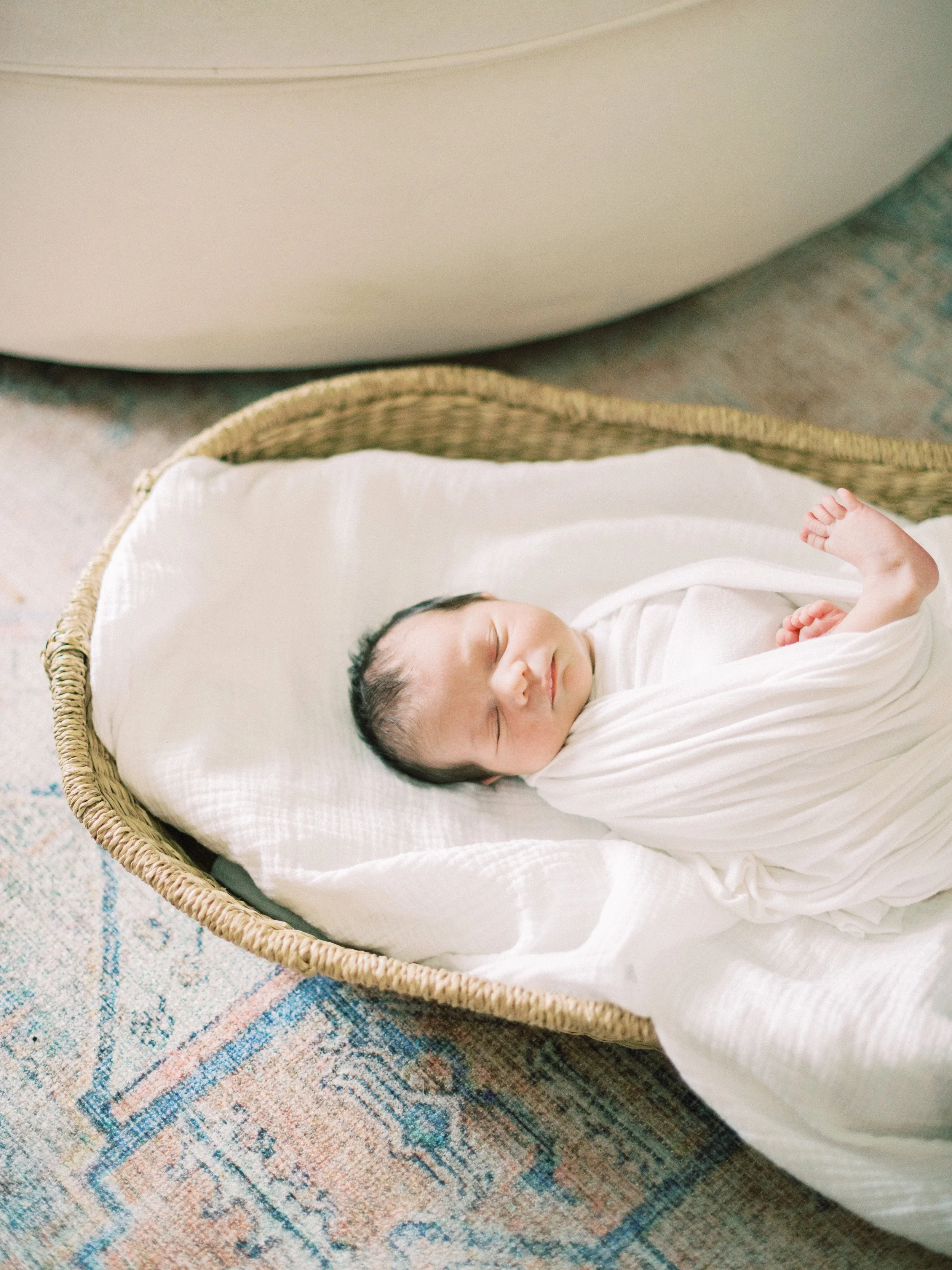A newborn baby sleeping in a wicker bassinet on a patterned rug.
