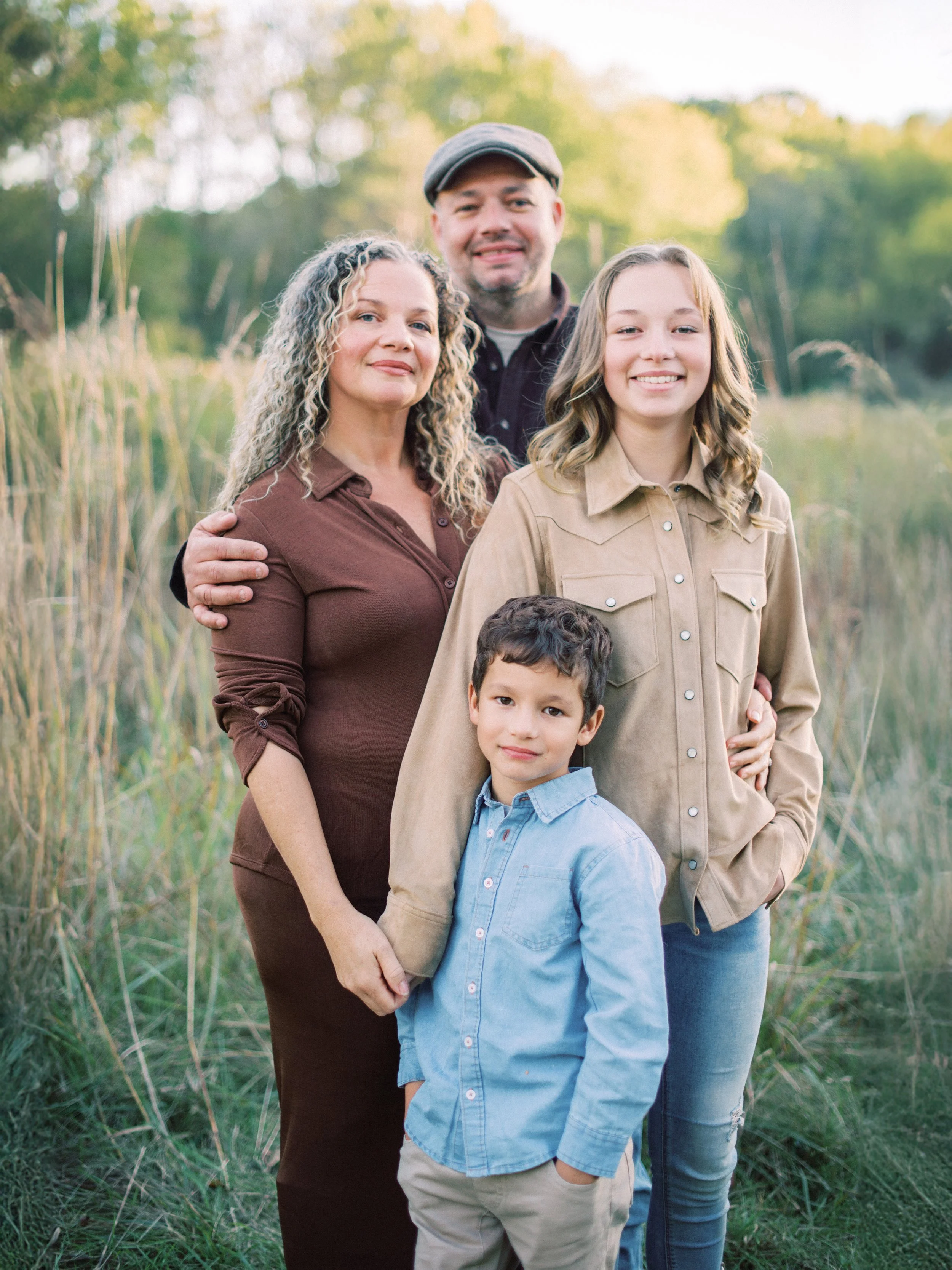 A family of five standing outdoors in a natural setting with trees and tall grass, smiling at the camera.