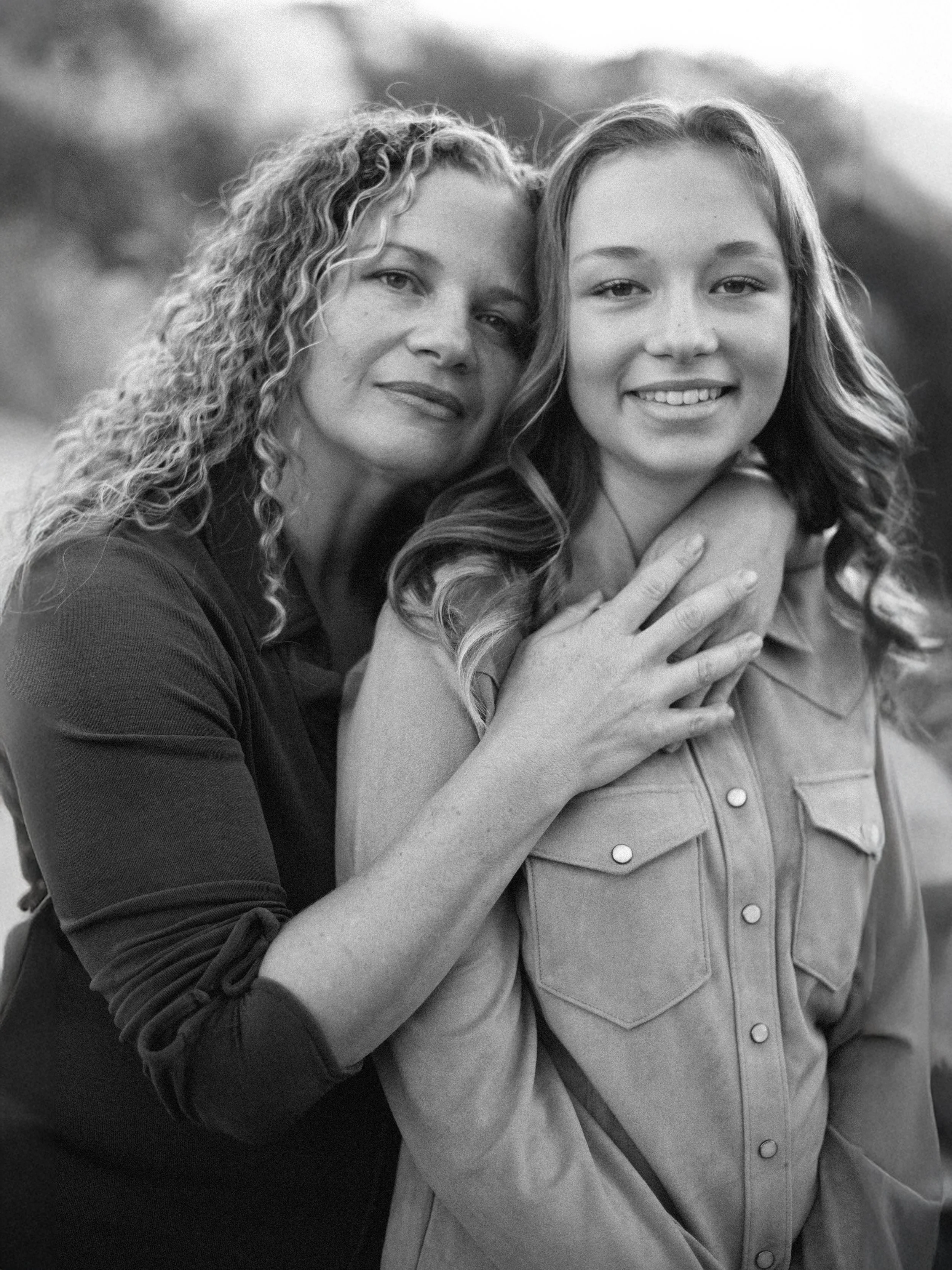 Black and white photo of a woman and a girl smiling, with the woman gently holding the girl from behind, both looking at the camera outdoors.