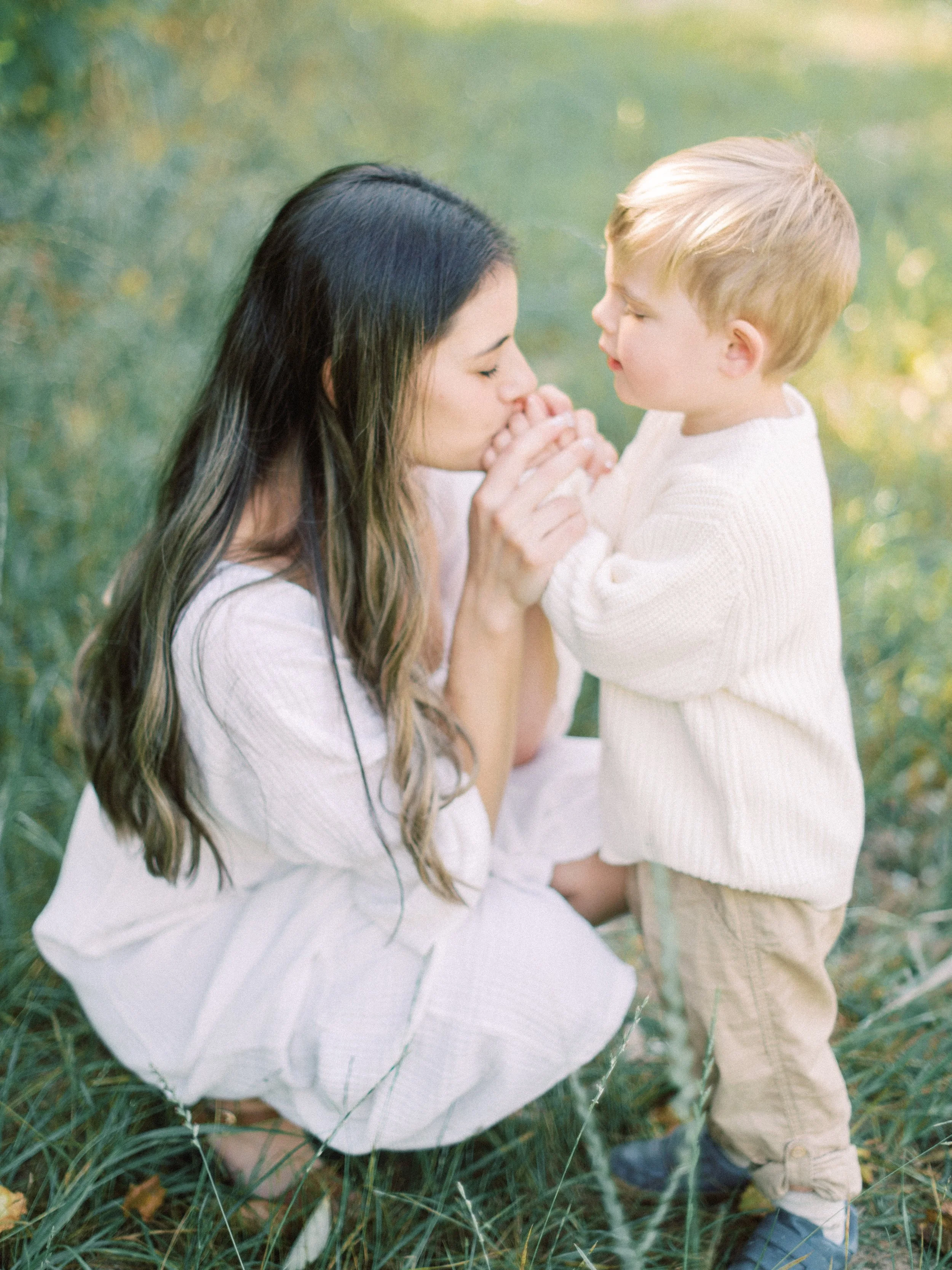 A woman crouching in a grassy field with a young boy, holding his hands and leaning in to touch noses, both dressed in white and beige clothing, surrounded by greenery.