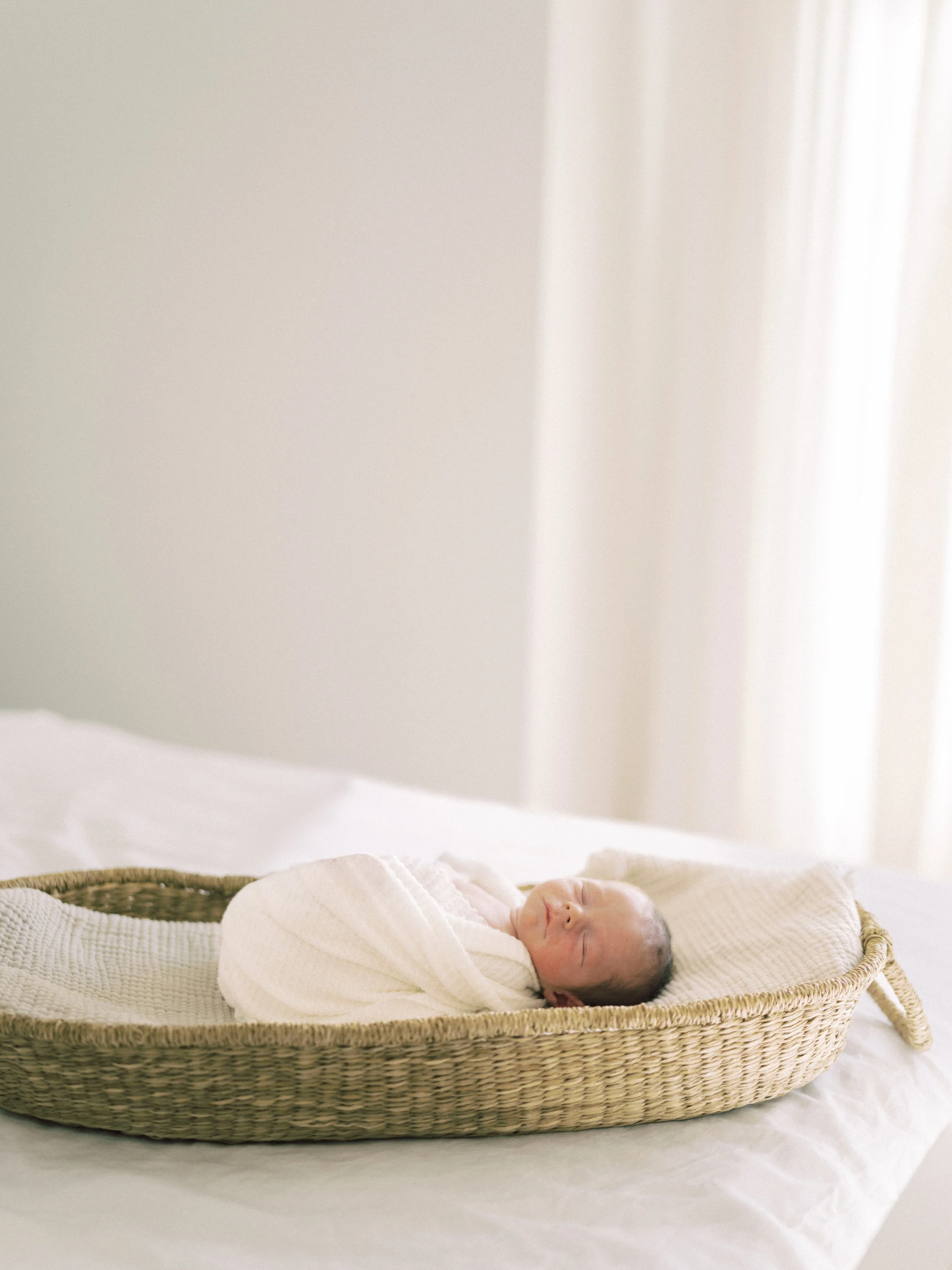 A newborn baby wrapped in a white blanket sleeping in a wicker bassinet on a white bed, with soft natural light coming through a curtain in the background.