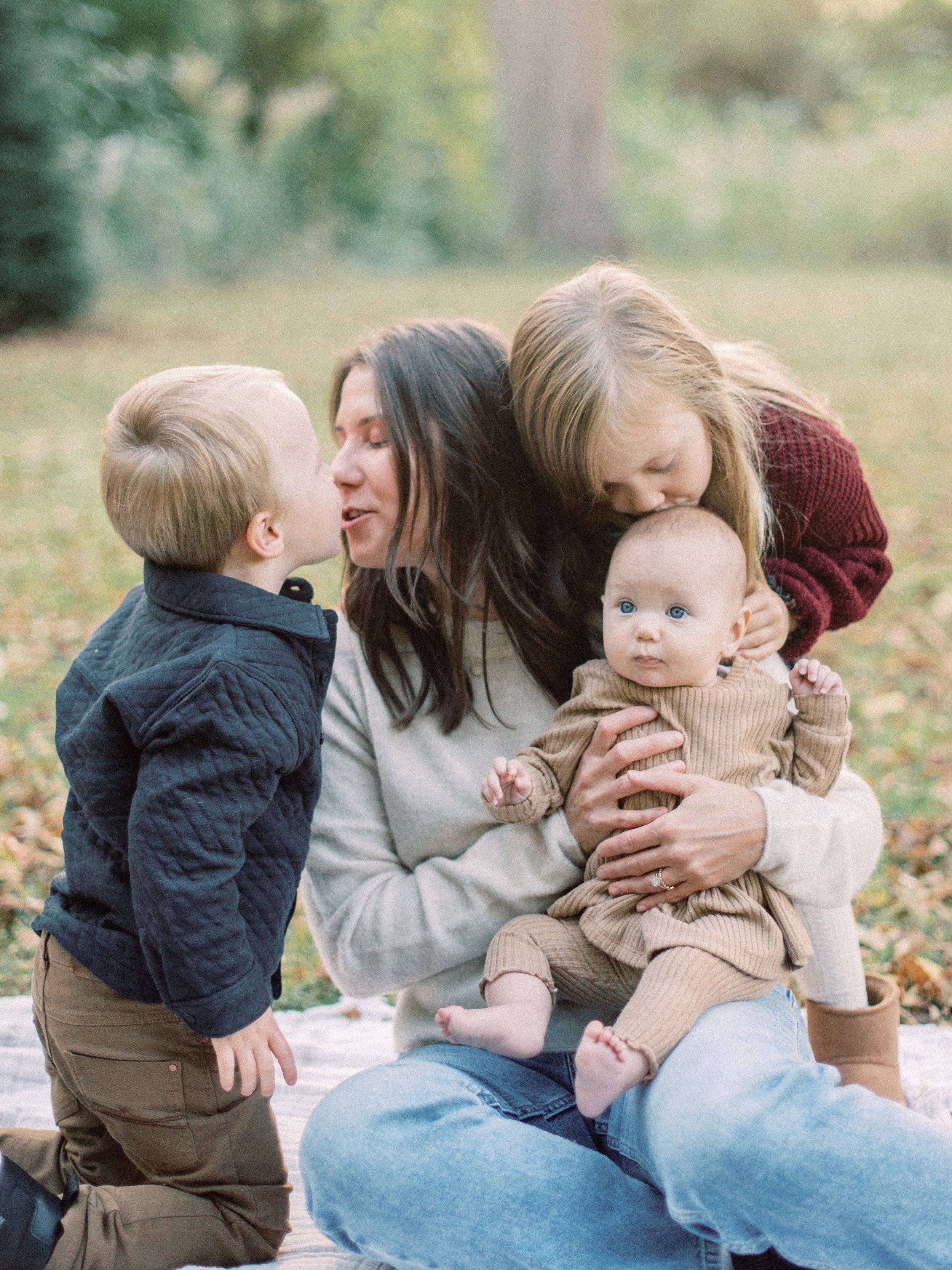 A woman with two children outdoors, one child is kissing her cheek, and the other is sitting on her lap, being kissed by an older girl.