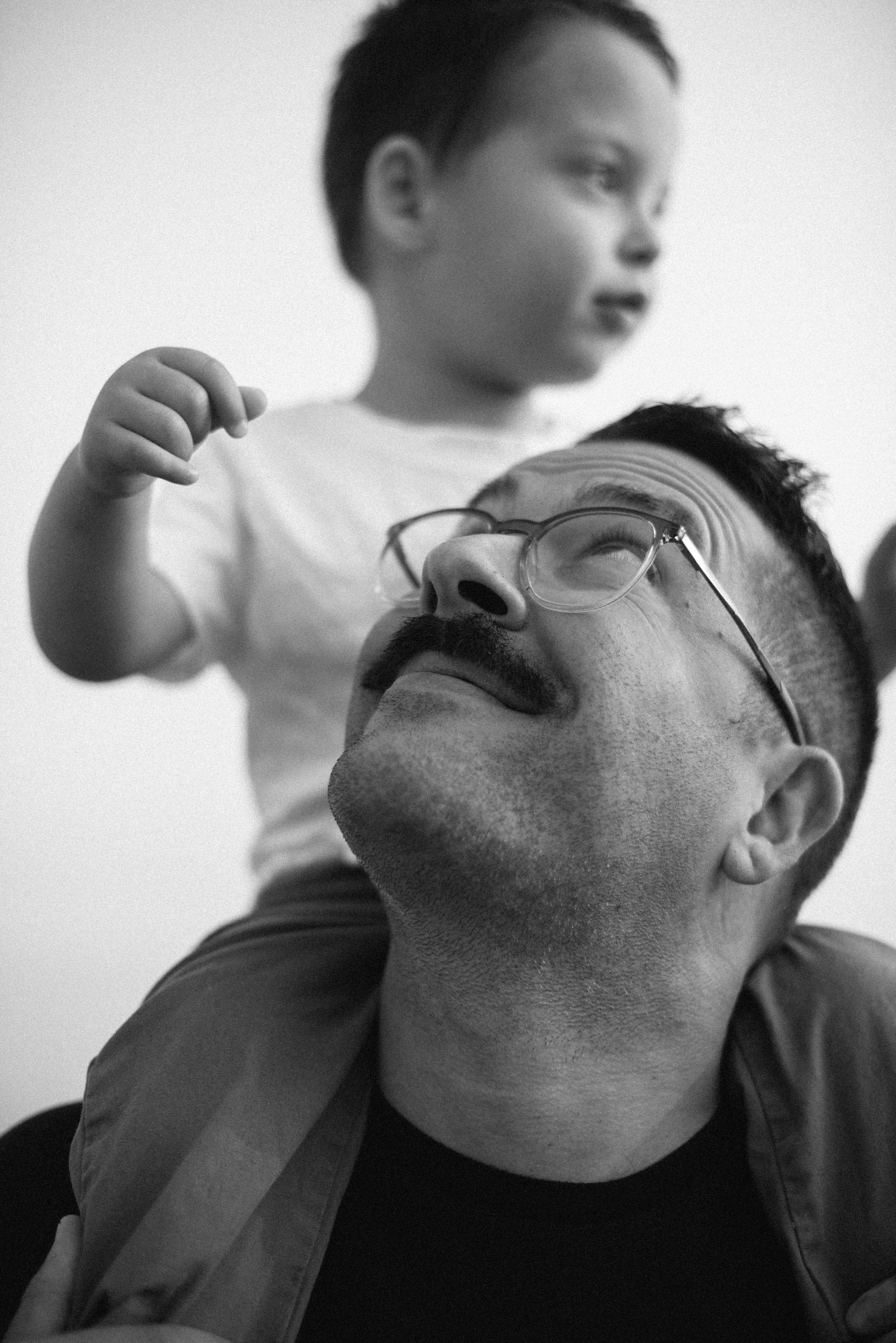A man with glasses and a mustache is carrying a young boy on his shoulders, both looking upward. The scene is in black and white.