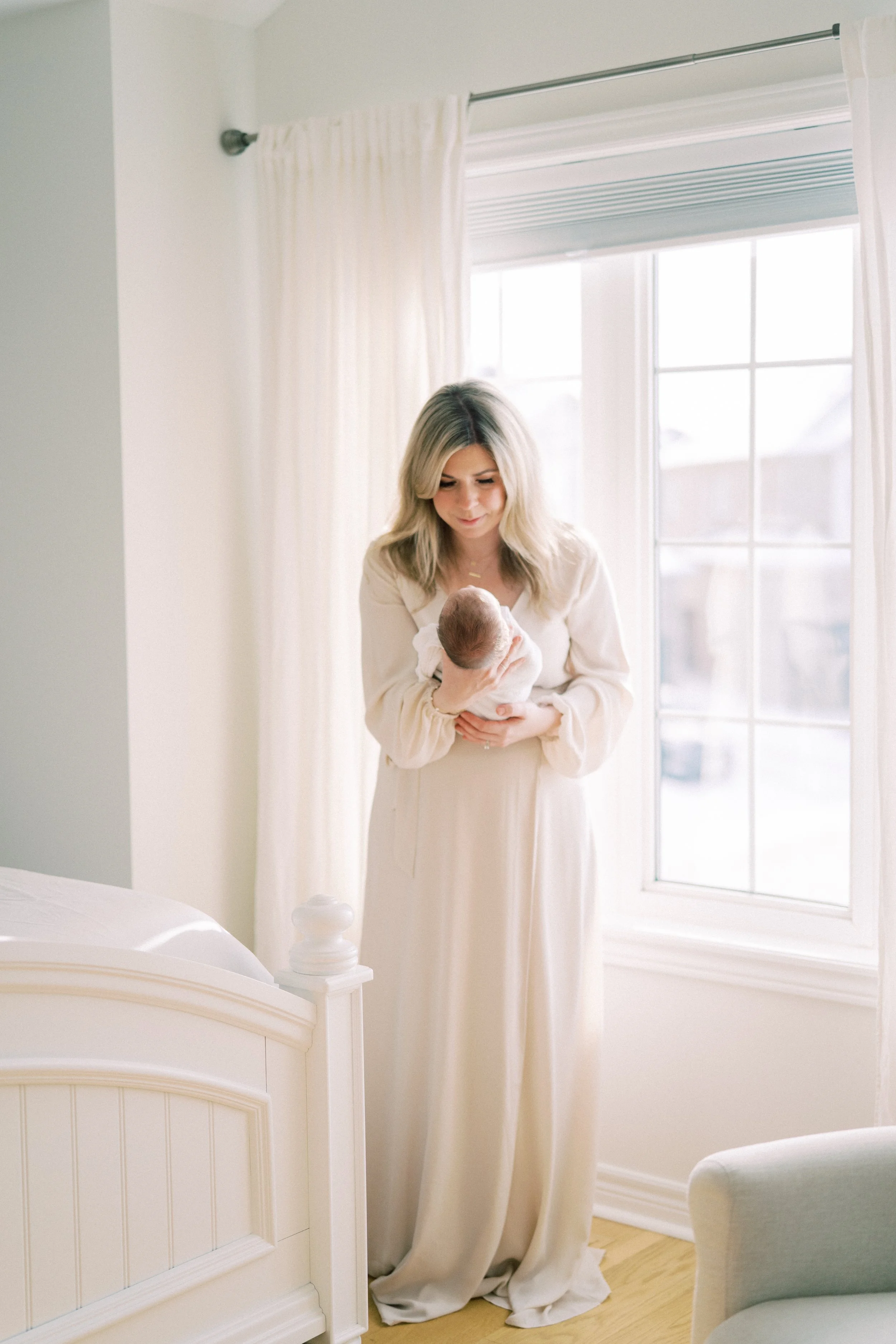 A woman in a long, cream-colored dress stands near a window, holding a newborn baby wrapped in a white blanket. The room has white curtains and a white bed, with soft natural light illuminating the scene.