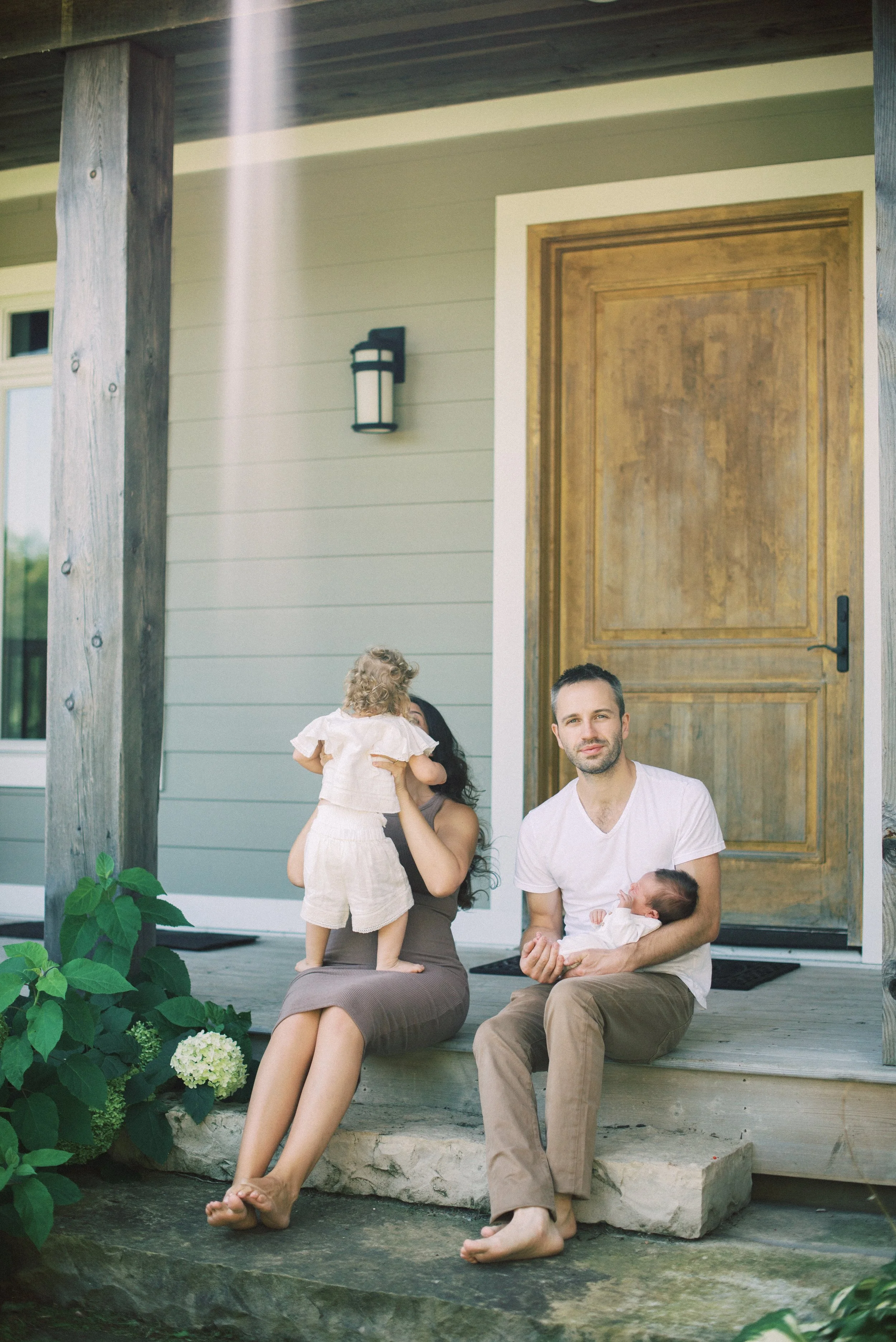 A family of four sitting on the front porch of a house, with a woman holding a toddler and a man holding a baby, outdoors on a sunny day.