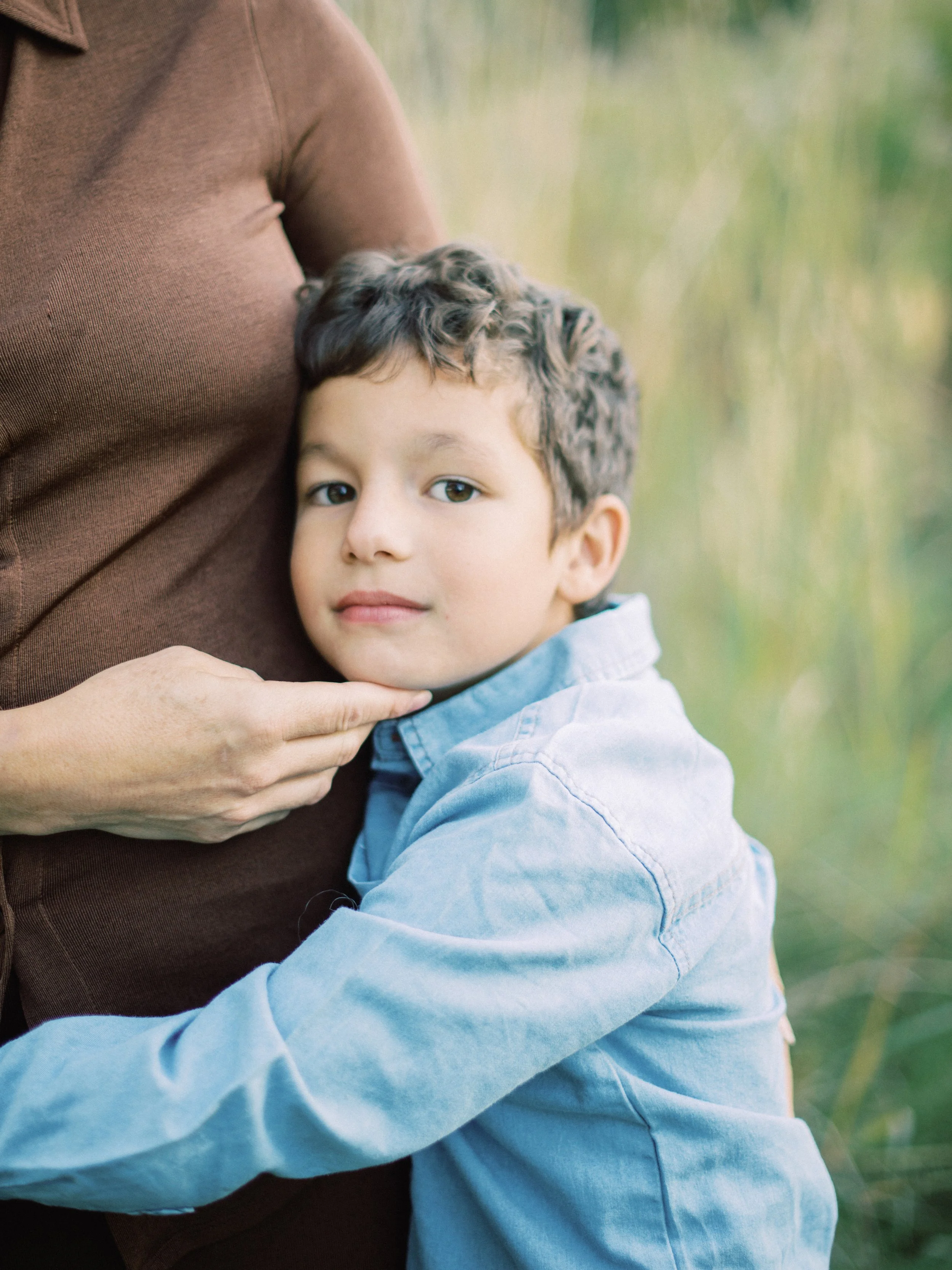 A boy leaning against an adult's chest, with the adult's hand gently touching his chin. They are outdoors with blurred green foliage in the background.