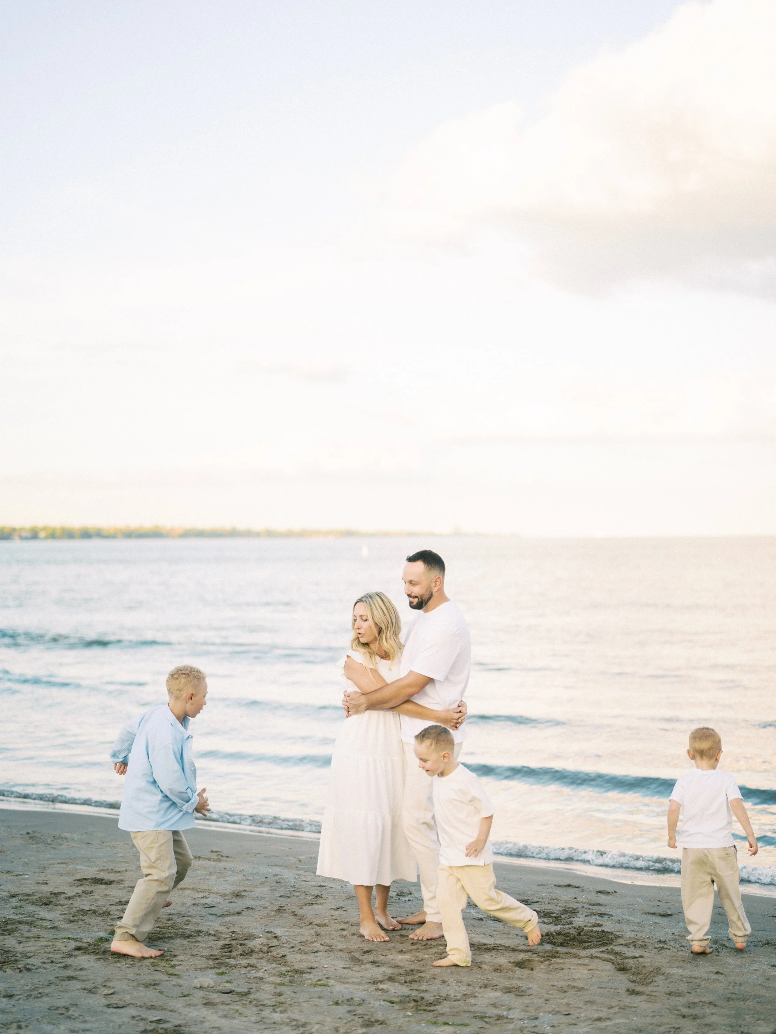 A family of five walking and playing on the beach near the water, with a cloudy sky above.