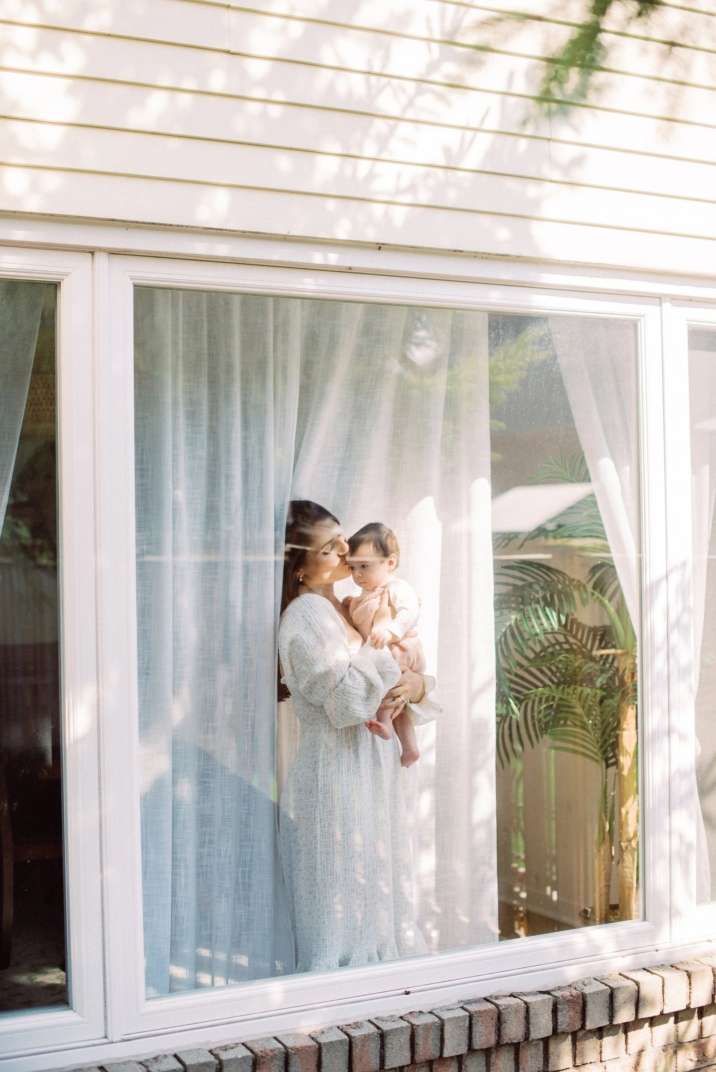 A woman holding a child inside, viewed through a large glass window with white curtains and a potted plant inside.