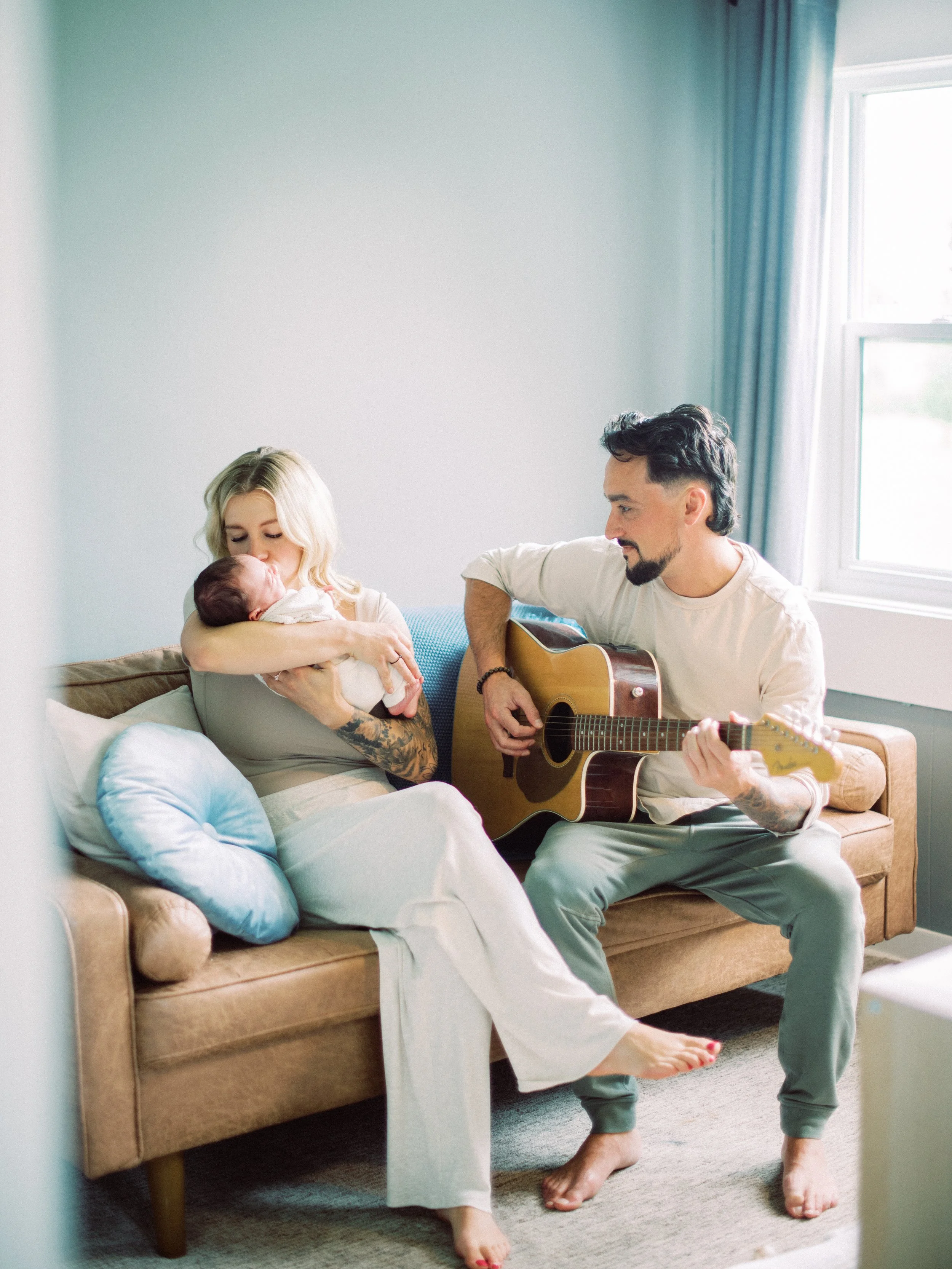 A woman holding a baby on a couch while a man plays guitar nearby in a bright living room.