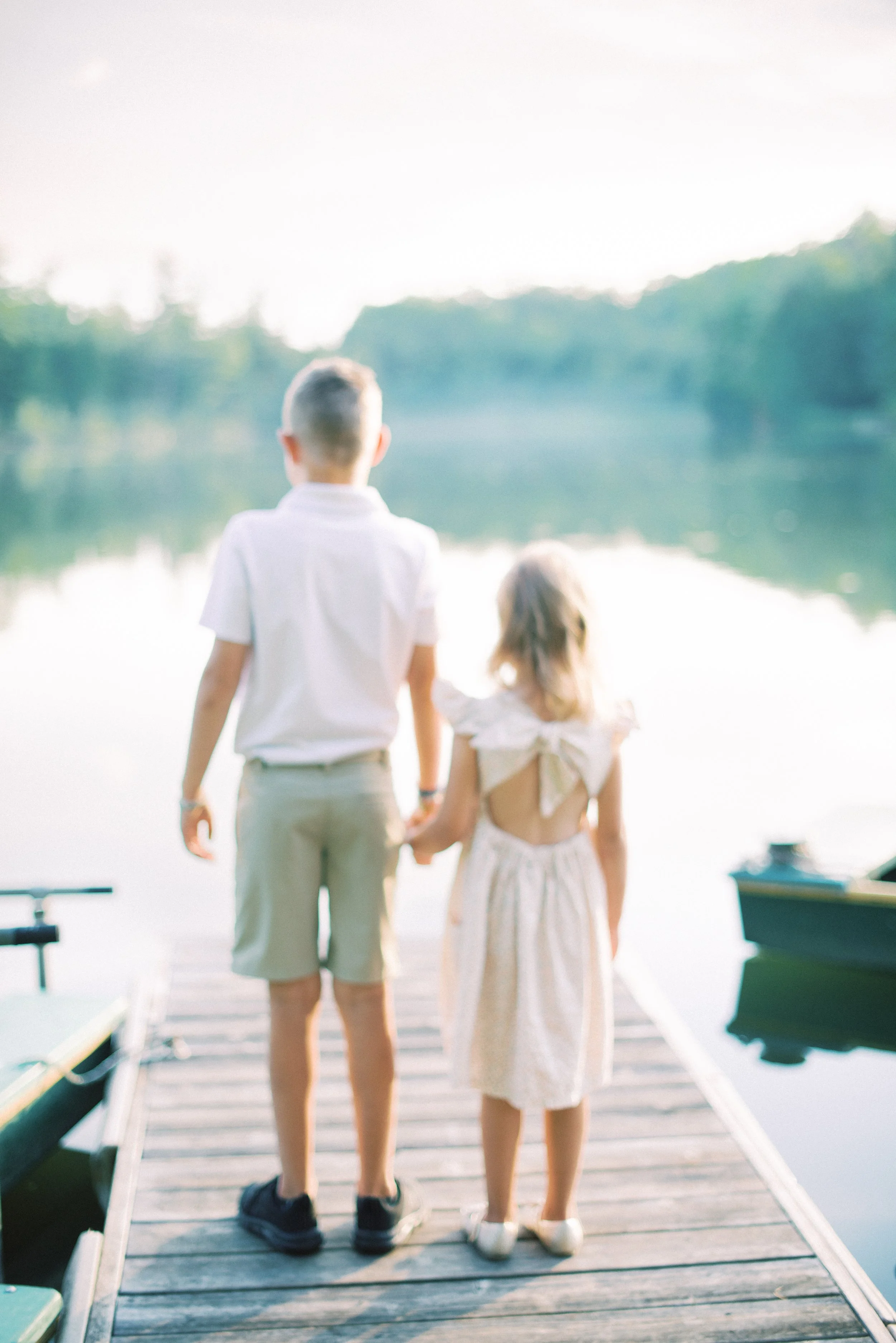 A boy and girl holding hands, standing on a dock by a lake, with boats on the water and trees in the background.