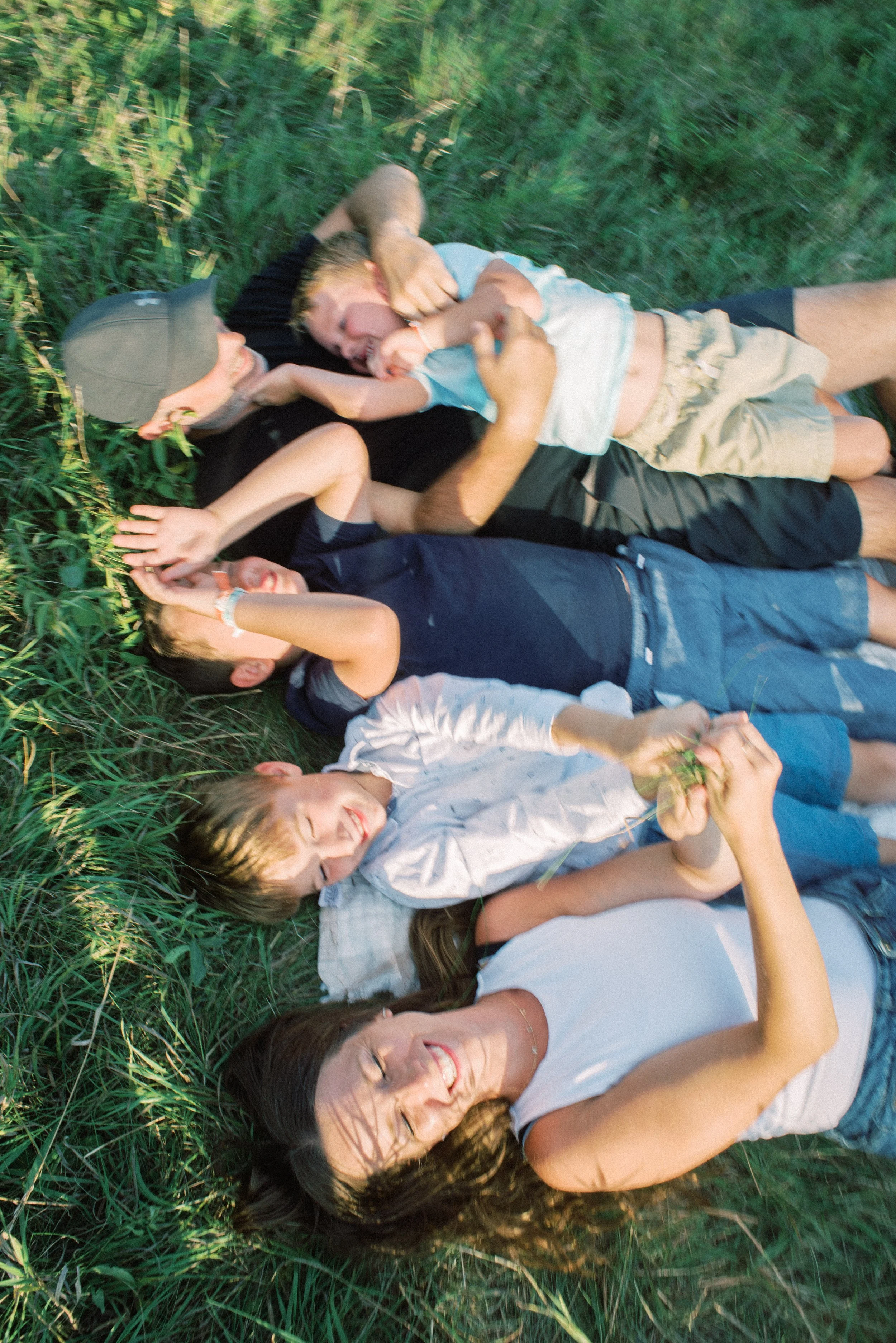 A woman and four children lying on grass, laughing and playing together during daytime.