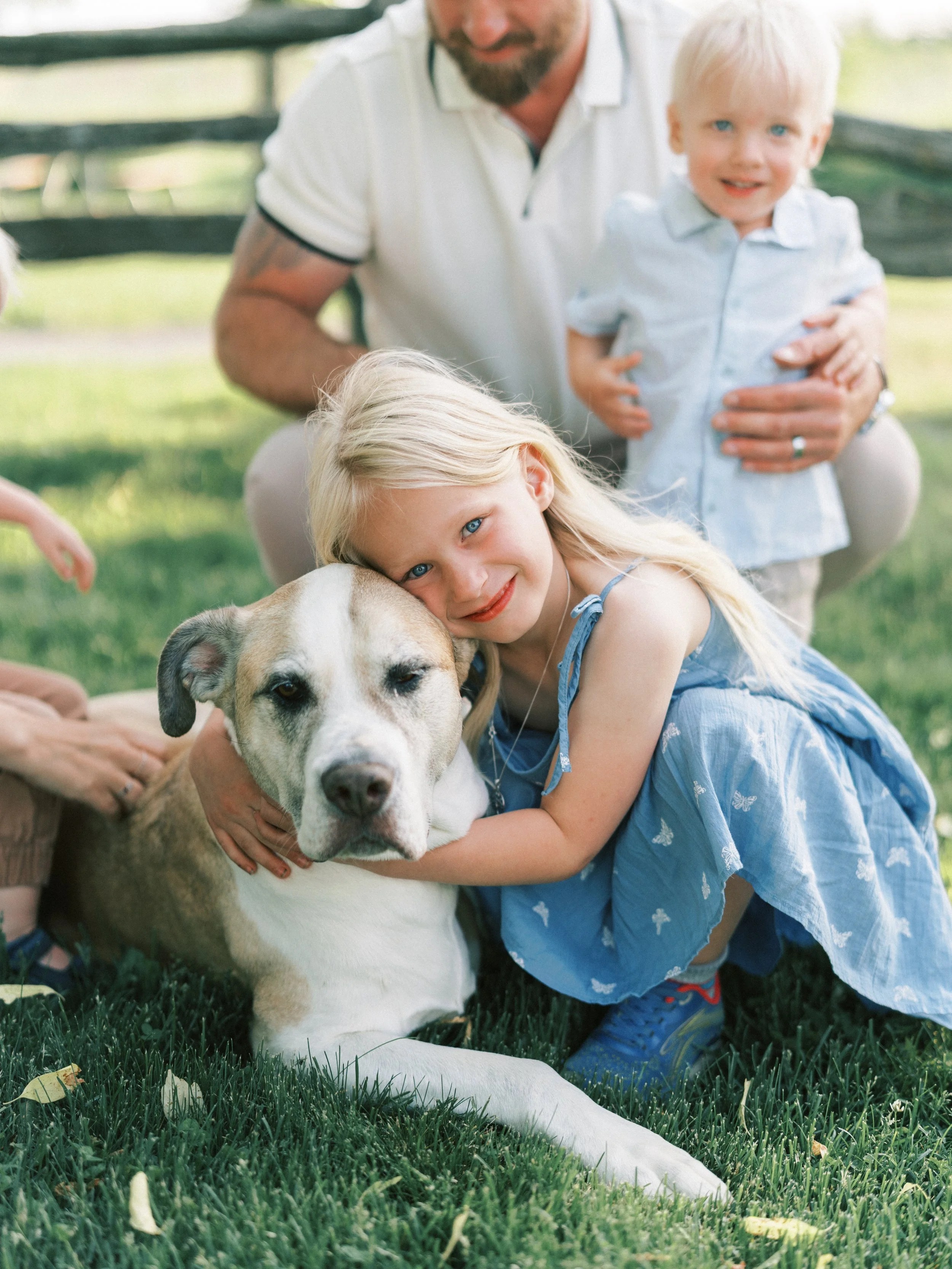 A young girl with blonde hair and blue eyes hugging a large dog lying on the grass, with a man and a young boy in the background on a sunny day.