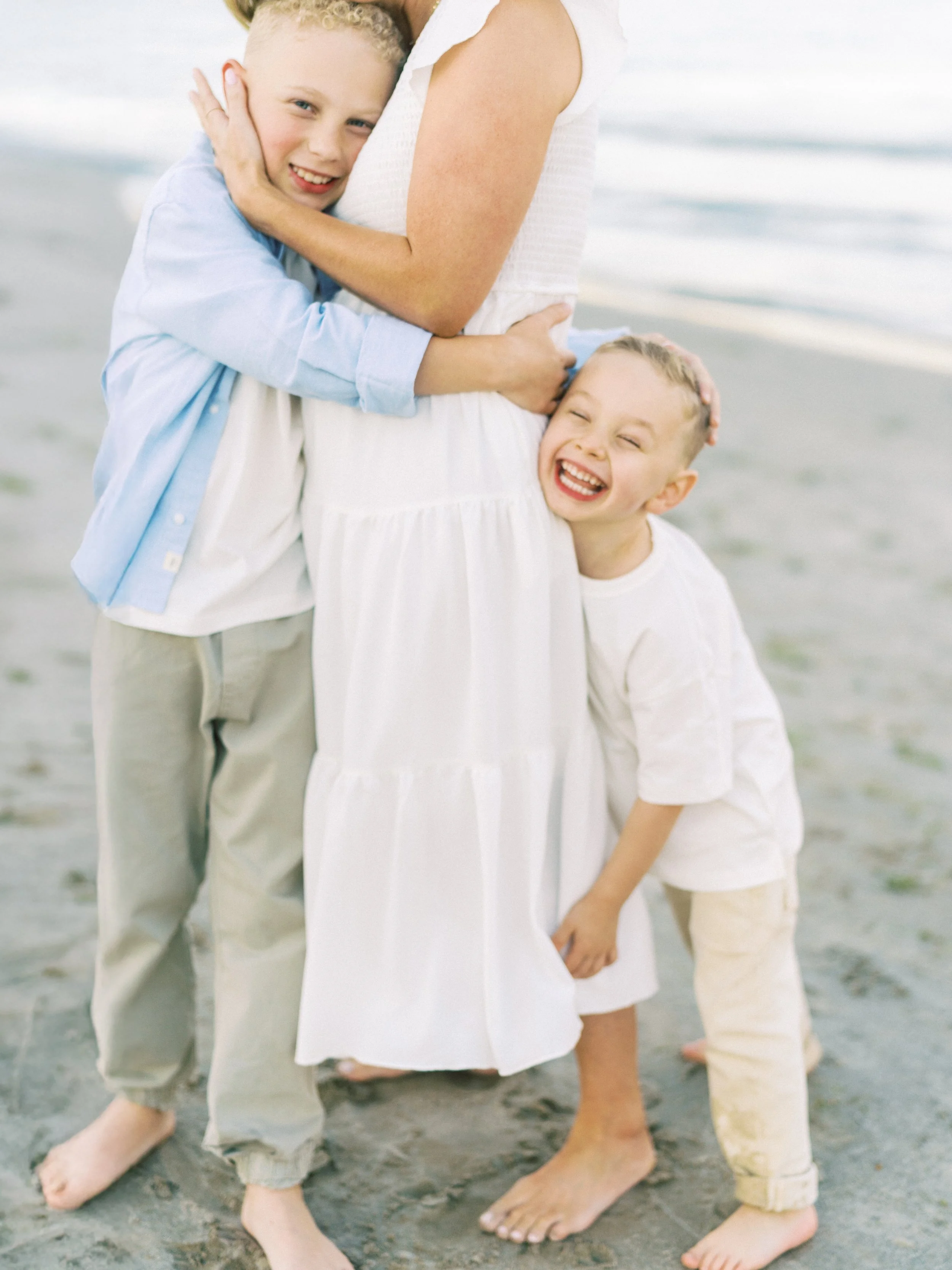 Two young boys hugging a pregnant woman on the beach, all smiling and barefoot.
