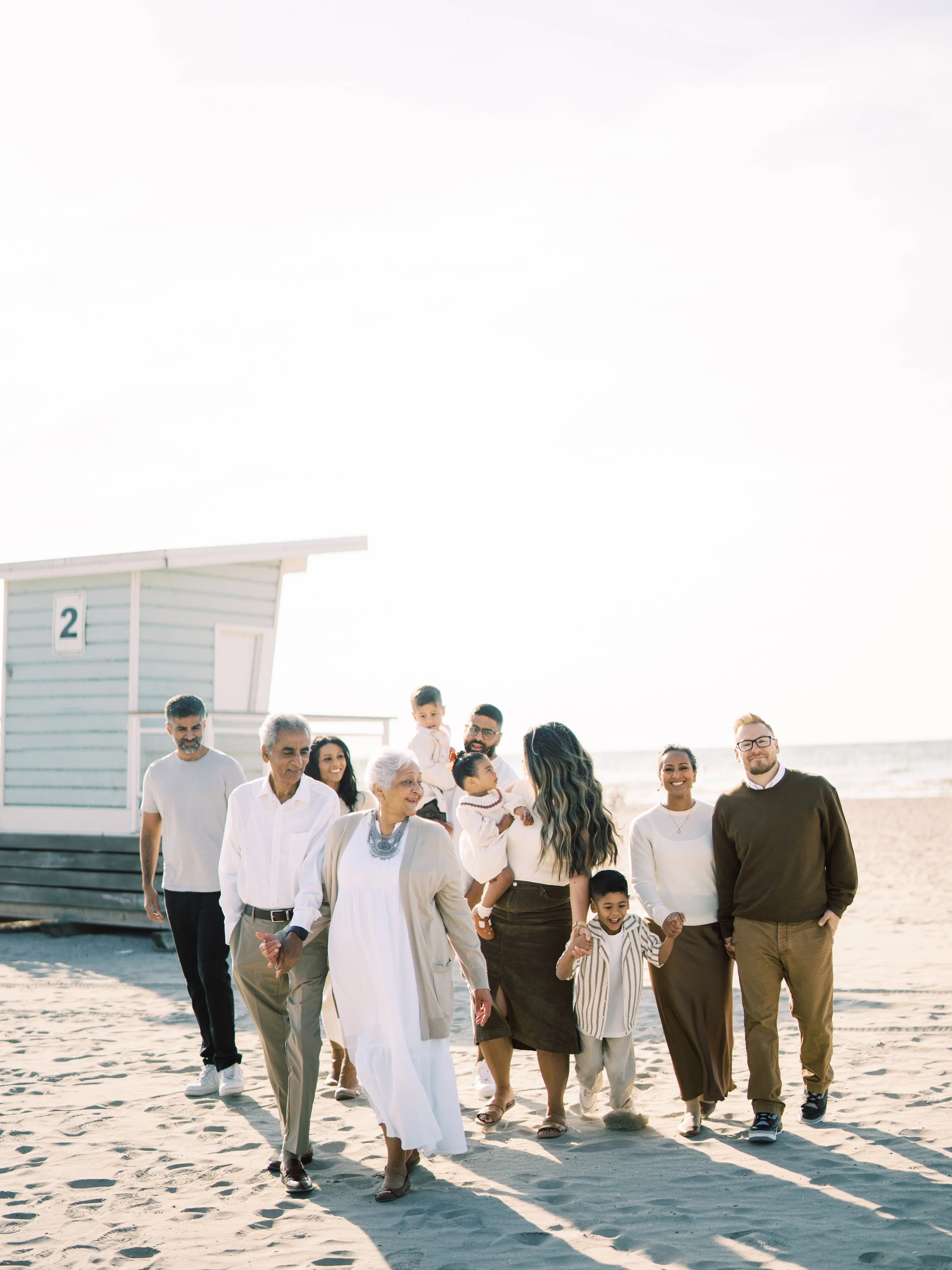 An extended family session, including children and adults, walking on Cobourg beach near a lifeguard hut, enjoying a sunny day.