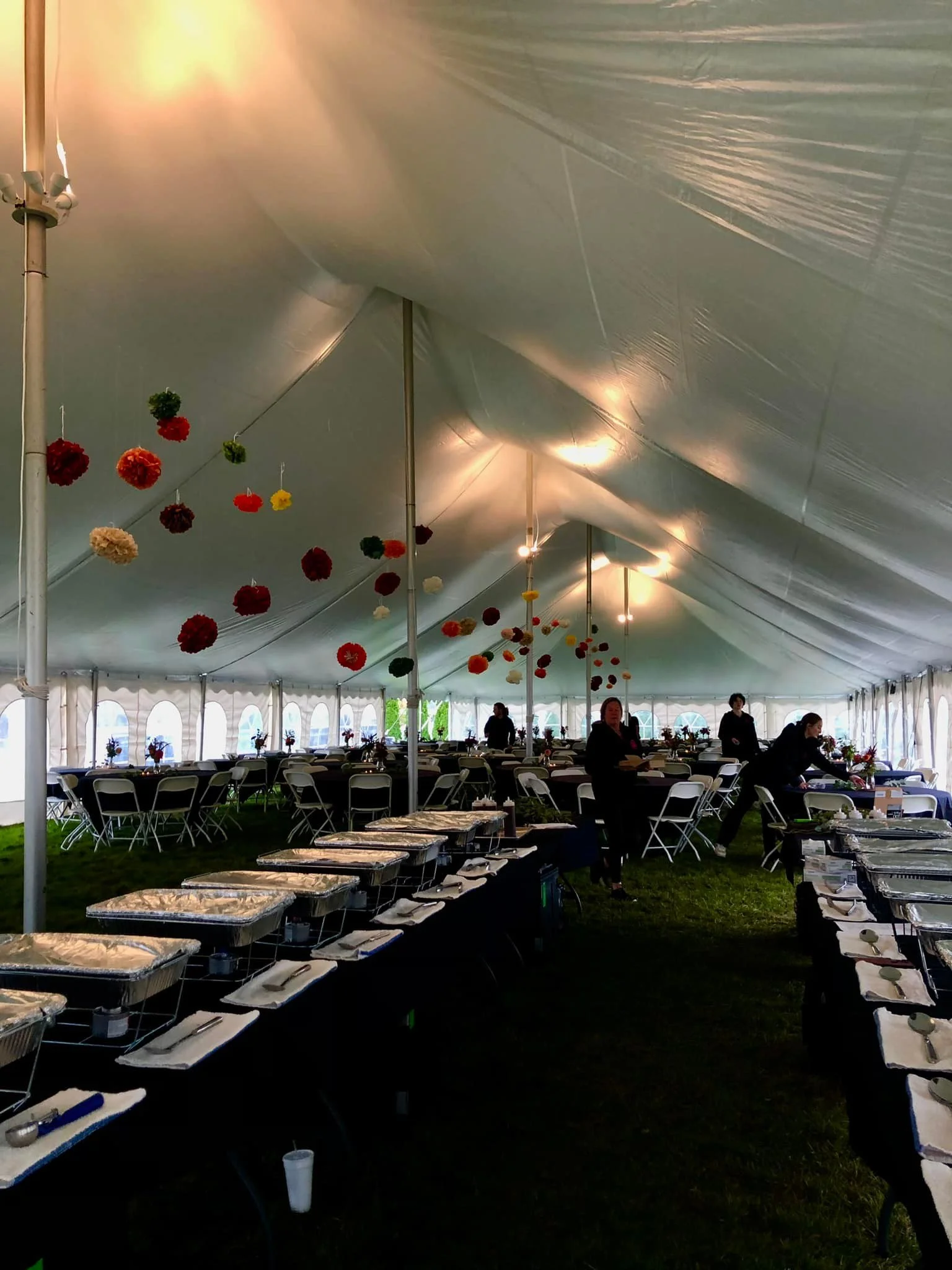 Large outdoor tent set up for an event with tables and chairs, food trays on serving tables, and decorative hanging pom-poms.