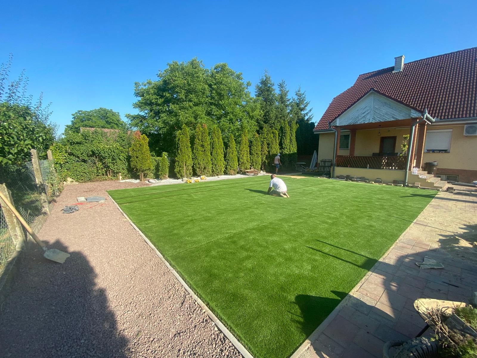Ein Haus mit einem roten Dach, umgeben von einem Garten mit frisch verlegtem Kunstrasen. Zwei Personen arbeiten im Garten, eine knieend auf dem Rasen. Im Hintergrund sind Bäume und Hecken zu sehen. Der Himmel ist klar und blau.