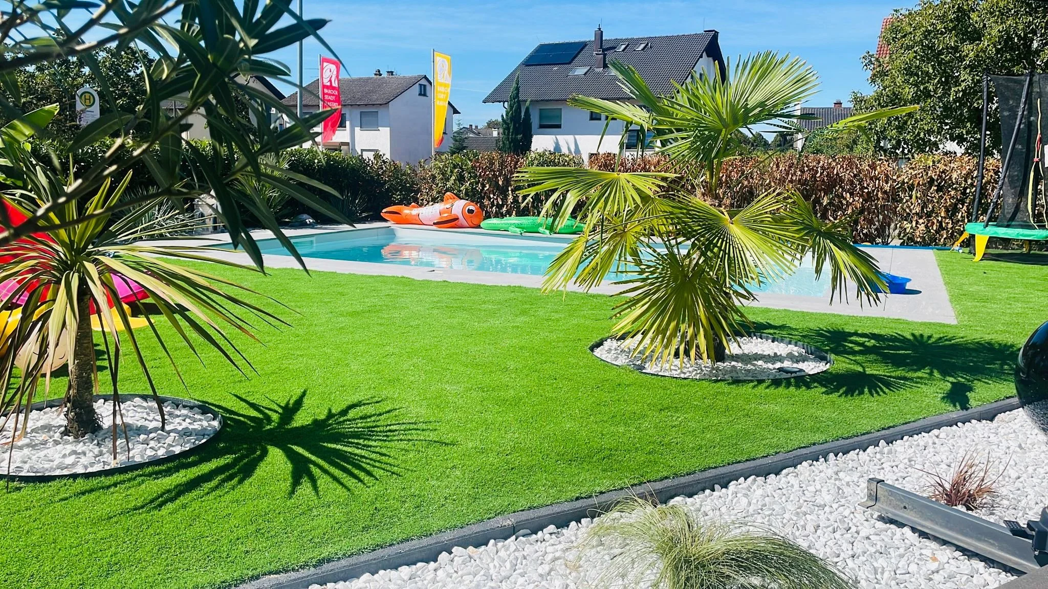 Garten mit einem kleinen Pool, Palmen und grüner Rasenfläche, umgeben von weißen Kieselsteinen. Im Pool liegen bunte aufblasbare Spielzeuge.