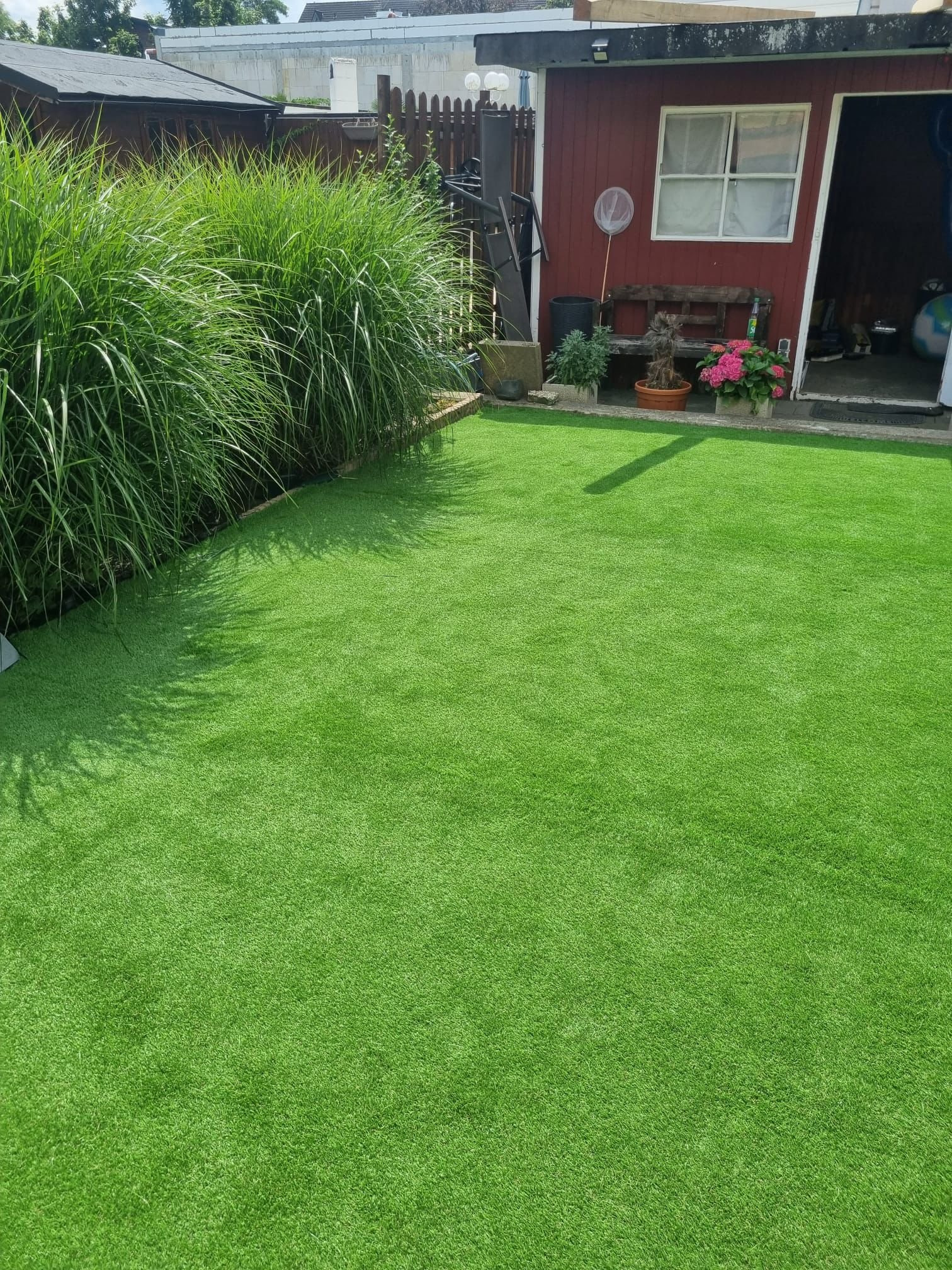 Hof mit grünem Kunstrasen, hohe Grasbüsche im Hintergrund, ein rotes Gartenhaus mit Fenster und offener Tür, bepflanzte Töpfe und Gartenmöbel daneben.