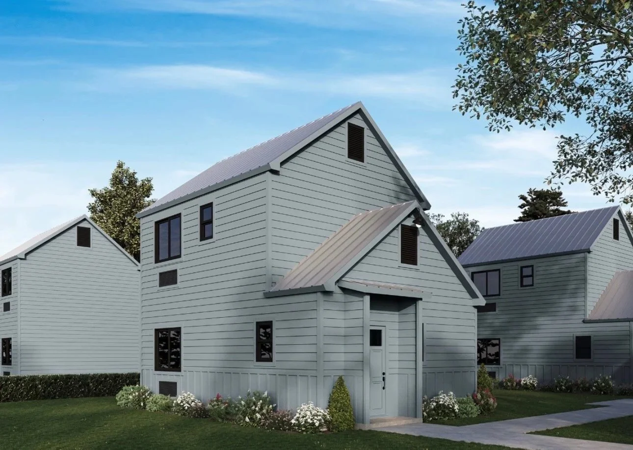 A row of modern, light gray houses with metal roofs, surrounded by green lawns and small flower gardens, under a partly cloudy sky.