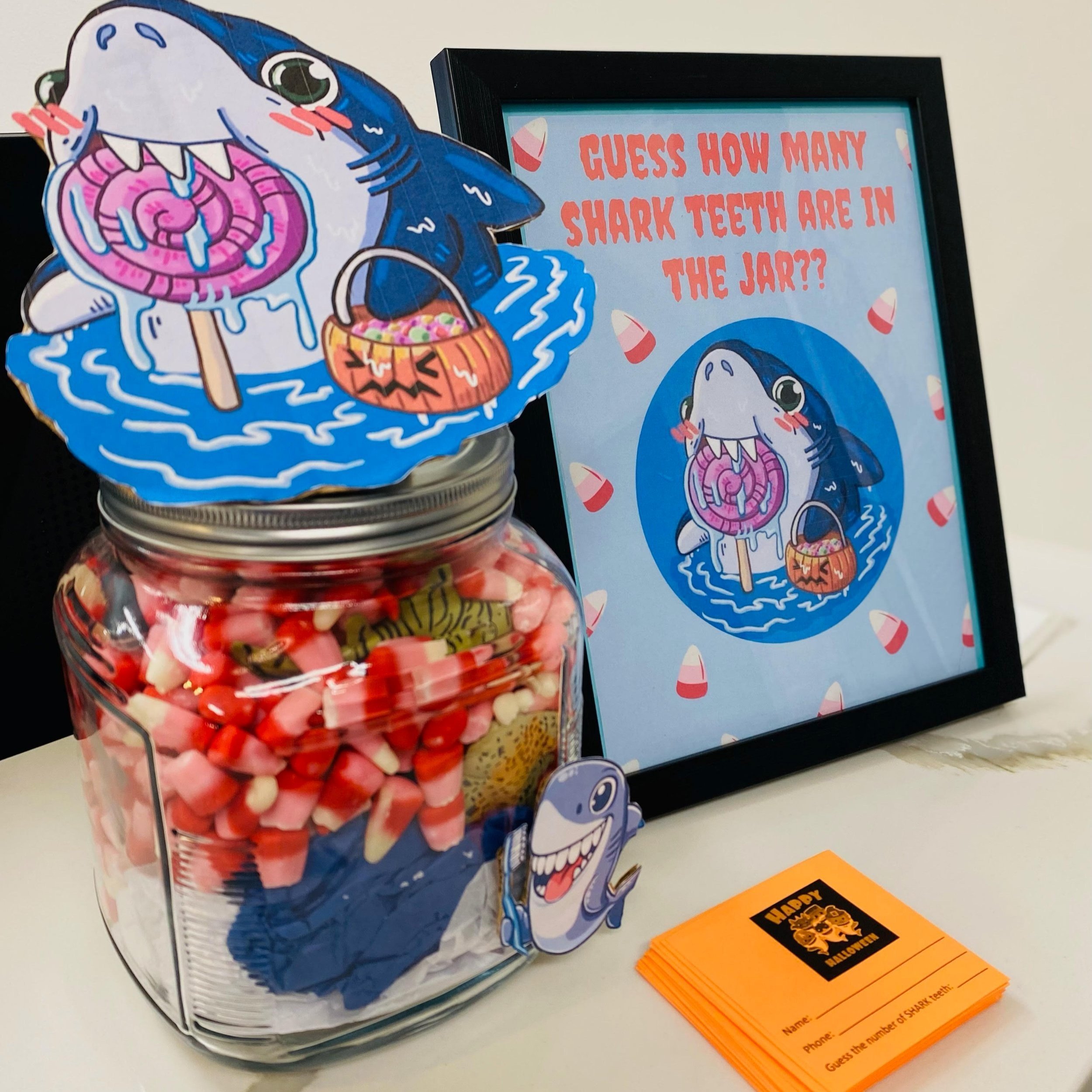 A jar filled with red and white candy, with a shark sticker on the front, sits next to a framed sign that says, "Guess how many shark teeth are in the jar?" The sign also has a picture of a cute cartoon shark with a lollipop and a pumpkin basket, alo