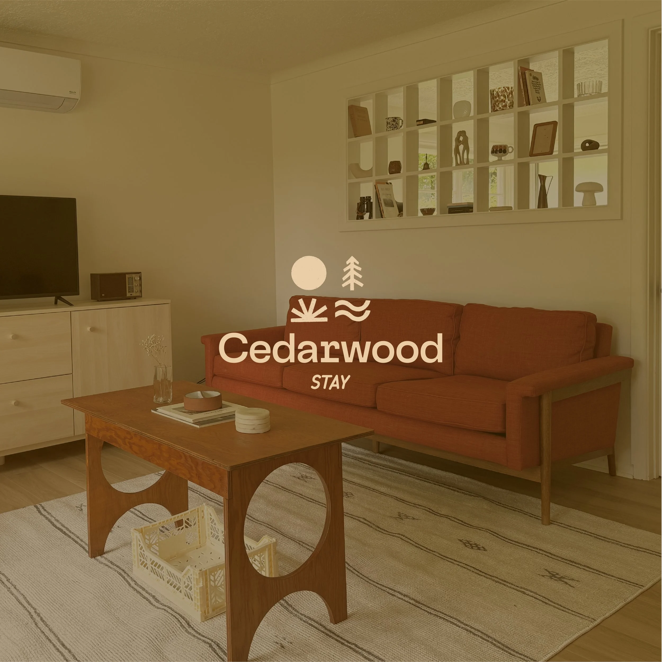 Living room with a red sofa, wooden coffee table, white dresser with a TV and radio, and a white wall shelf filled with decor items and books. The room features a beige area rug, a glass vase, and minimalist decor.