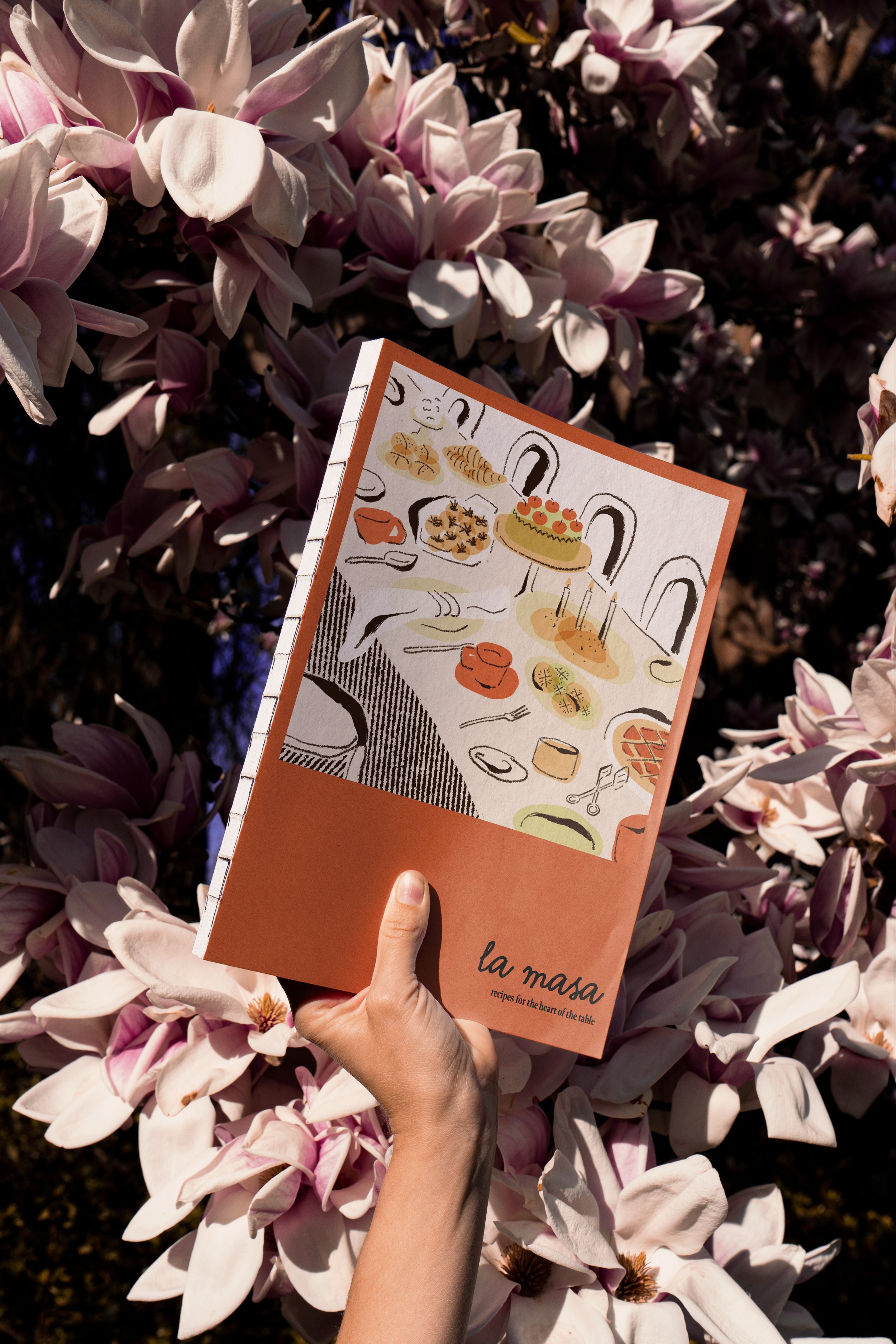 A person holding a cookbook titled 'la masa' in front of pink magnolia flowers. The cover features an illustration of a table set with various dishes and tableware.