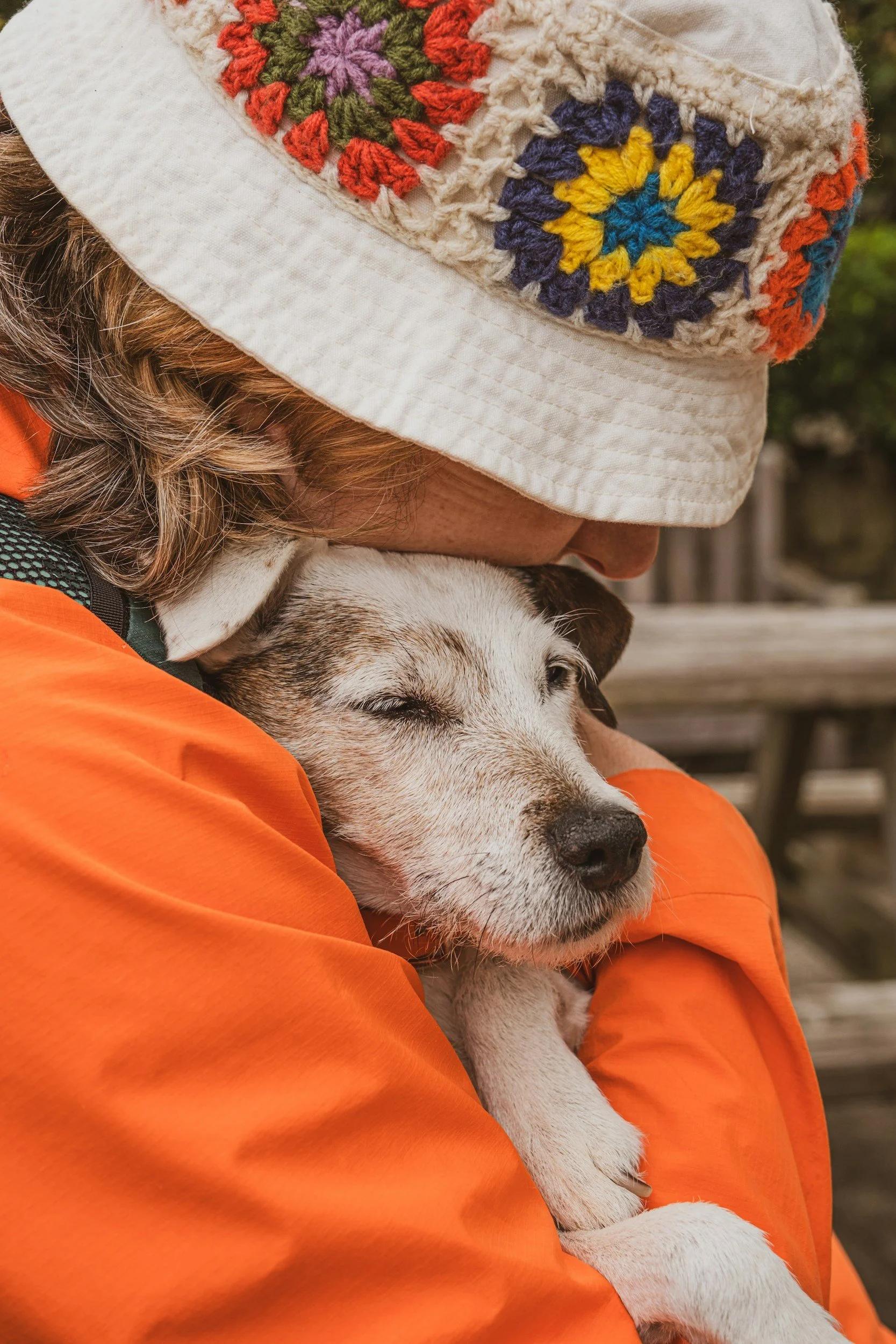 A woman with a hat wearing an orange jacket is holding a dog with its eyes closed.