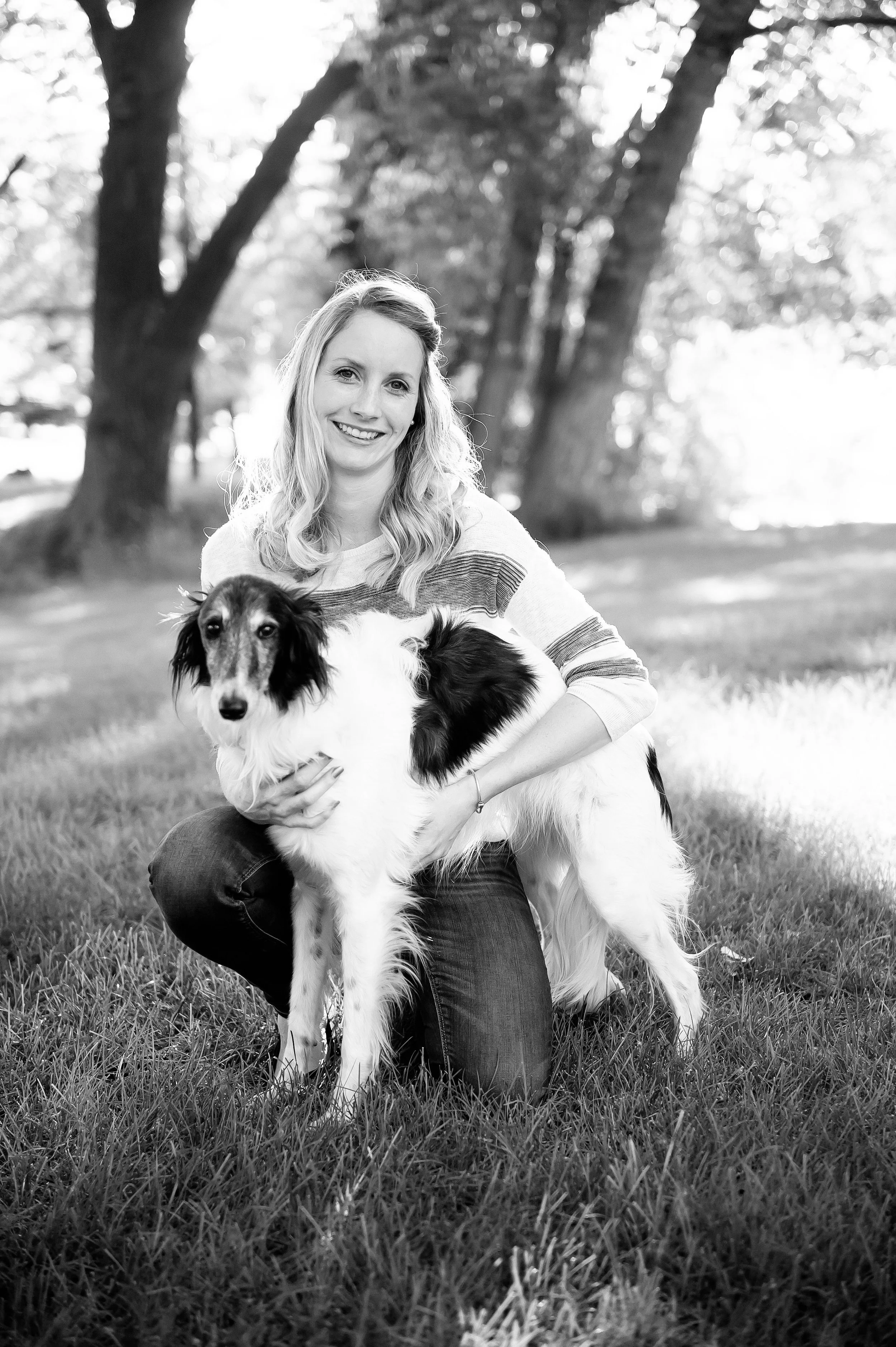 A woman with long hair sitting on the grass and holding a large dog with long, dark ears and a white and black coat, outdoors with trees and sunlight in the background.