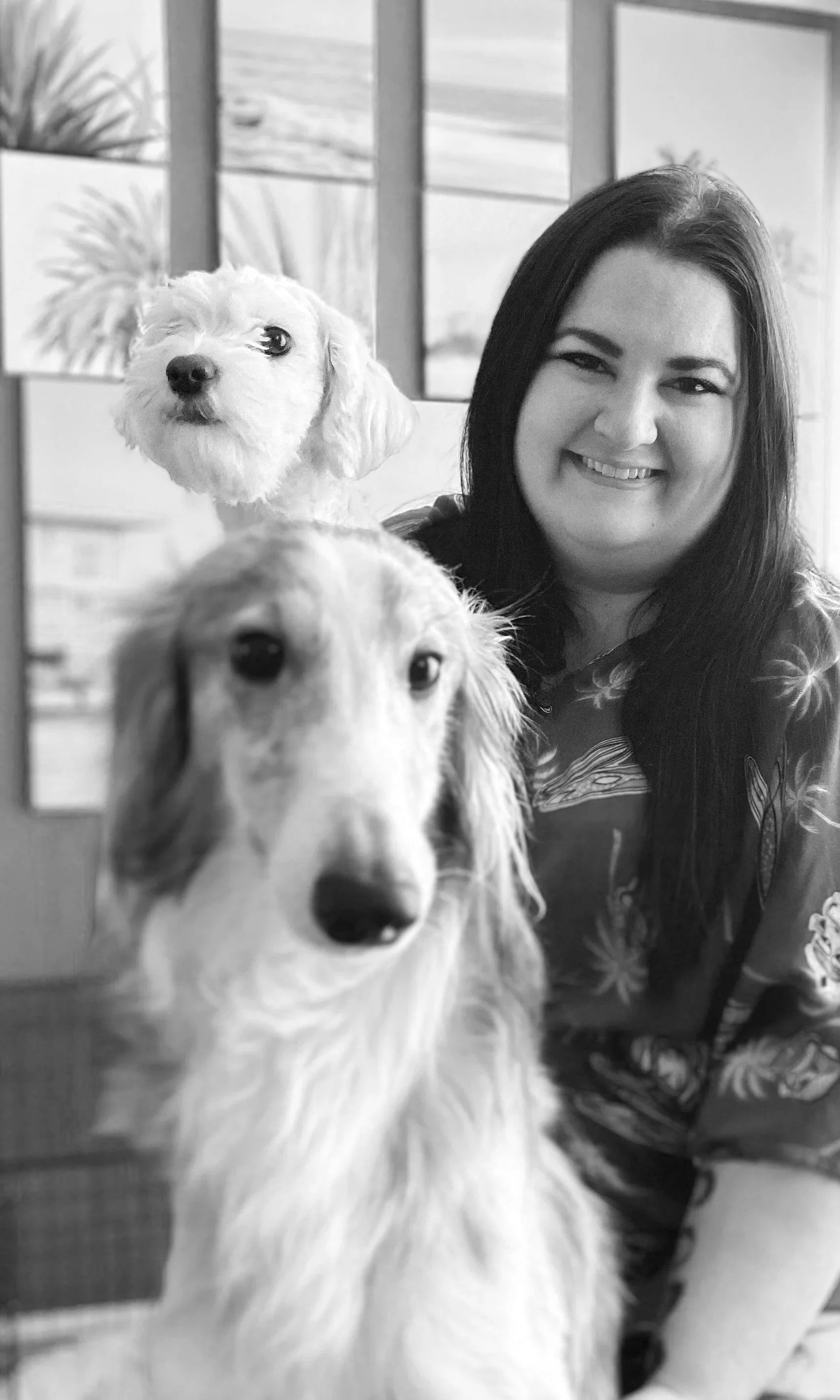 A woman smiling with two dogs, a small fluffy dog on her left and a long-haired dachshund in front of her, inside a house with windows in the background.