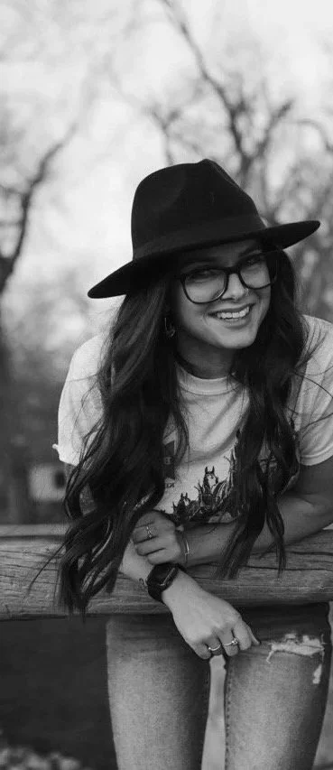 A smiling woman wearing glasses, a wide-brimmed hat, and casual clothes, leaning on a wooden railing outdoors.