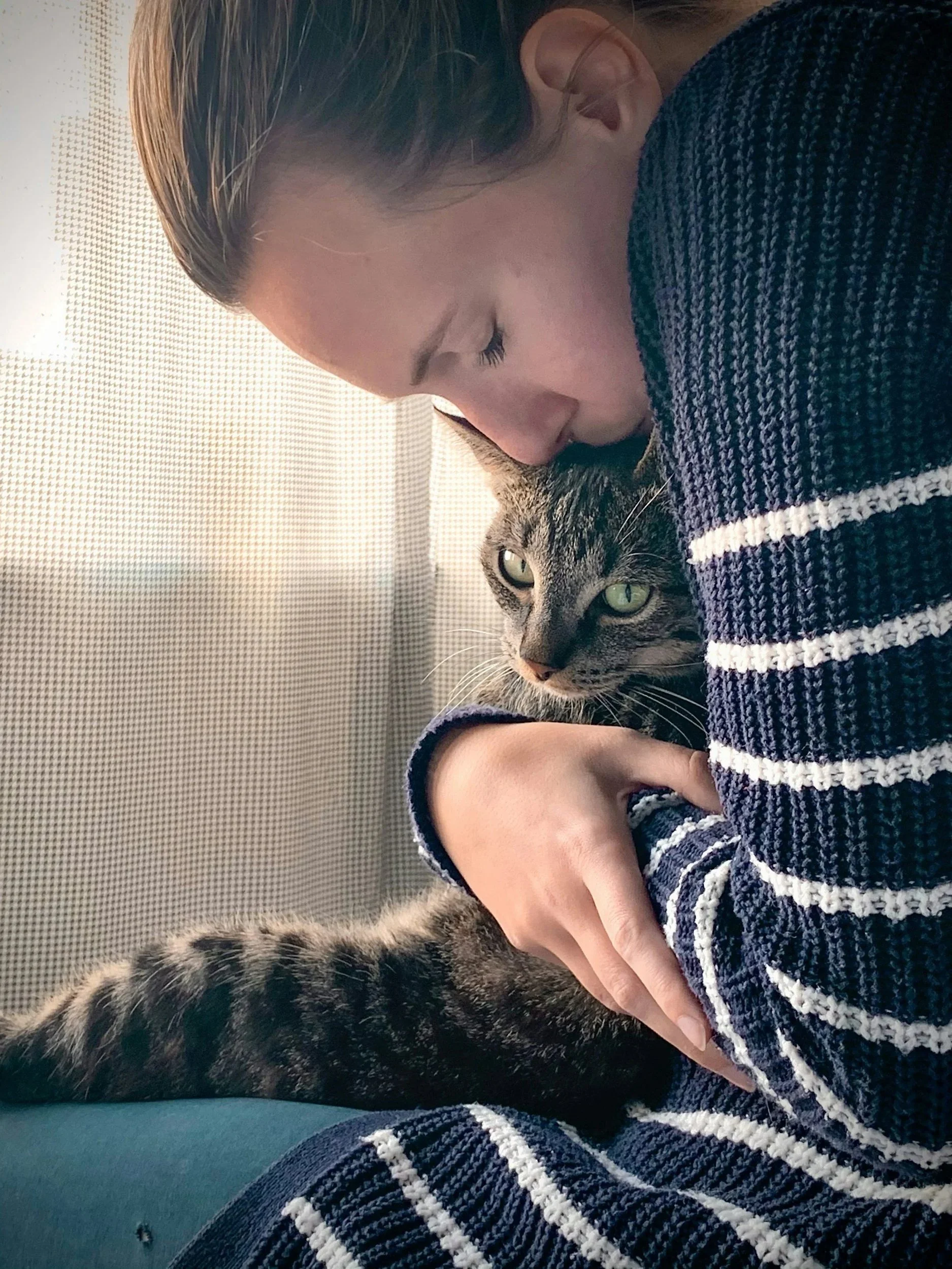 A person with closed eyes, wearing a striped sweater, gently holding a tabby cat with green eyes and striped fur.