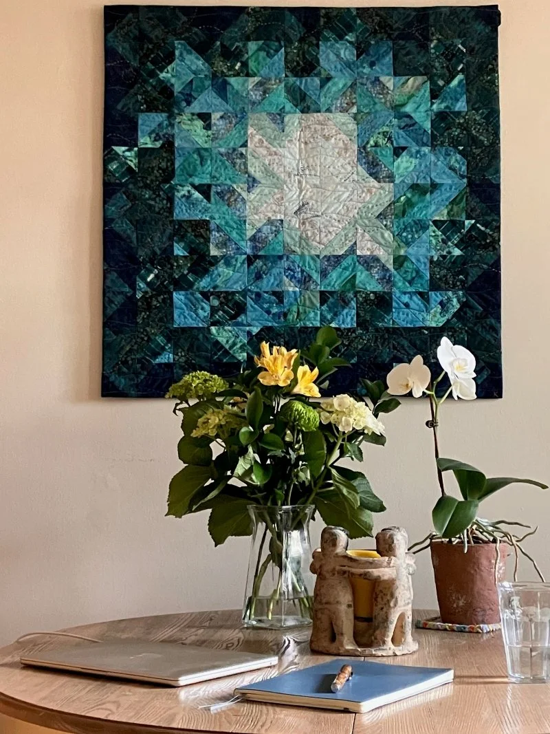 Wooden table with a closed MacBook Air, notebook, pen, vase of flowers, and white orchid in front of a blue geometric wall quilt