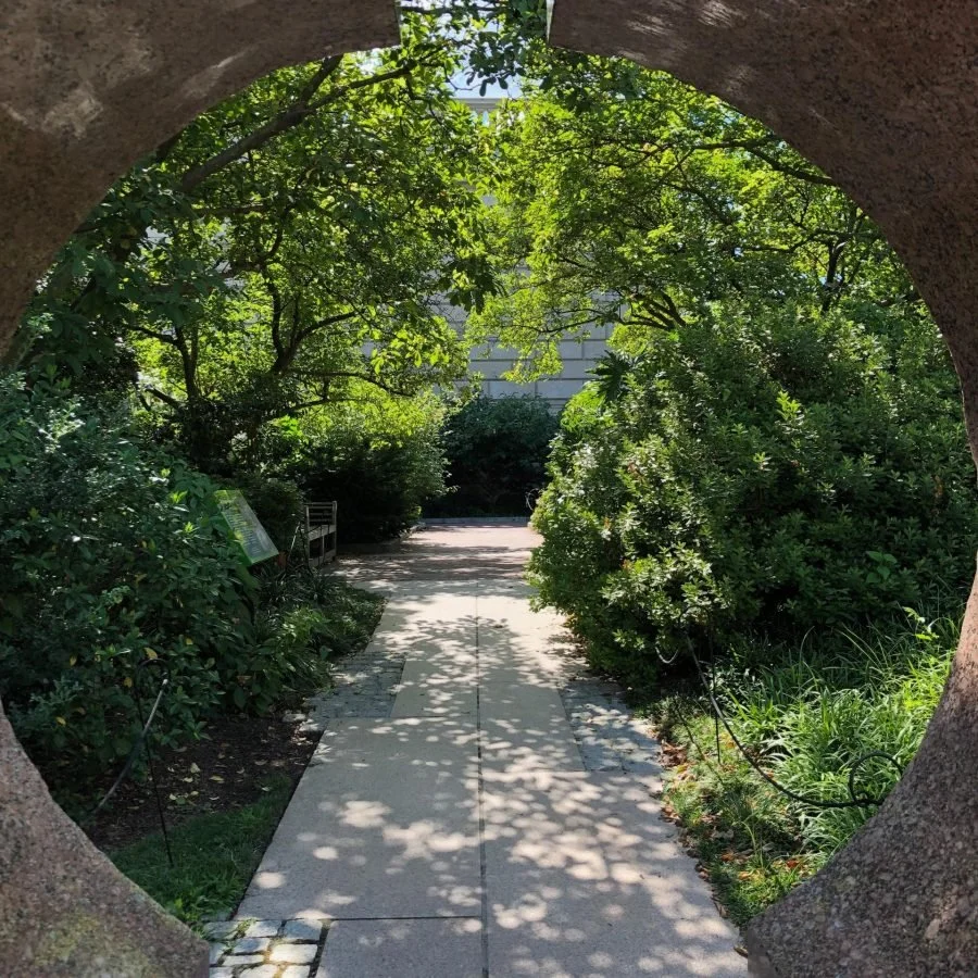 Circular stone arch framing a sunlit garden path lined with green shrubs and trees