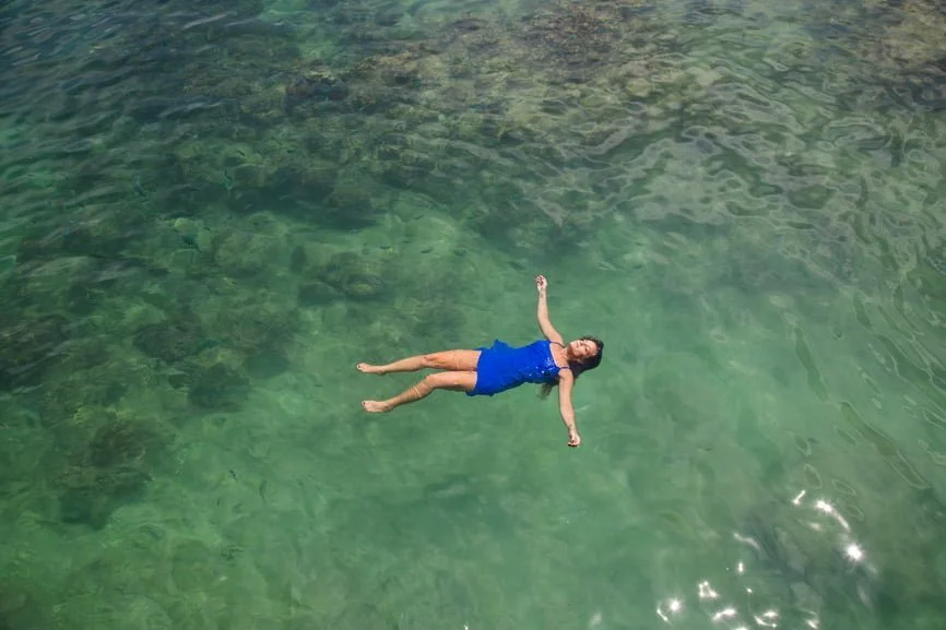 Person floating on their back in clear turquoise water wearing a blue swimsuit, viewed from above
