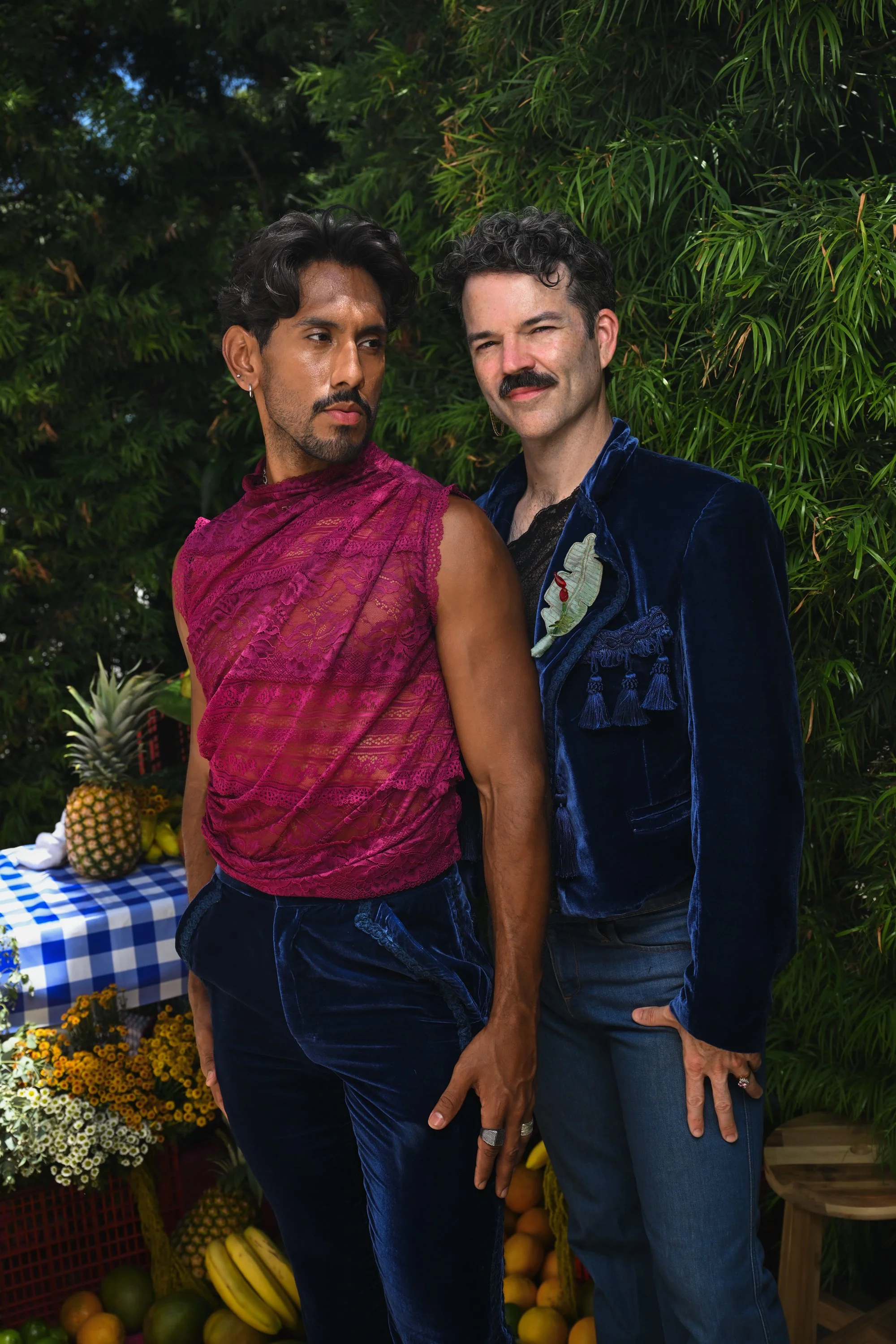 Two men standing close together outdoors near lush green foliage. One man with dark hair and a mustache wearing a blue velvet jacket, the other with short curly hair and a beard, wearing a pink lace top. There is a table with pineapples and bananas, and a display of various flowers in the background.