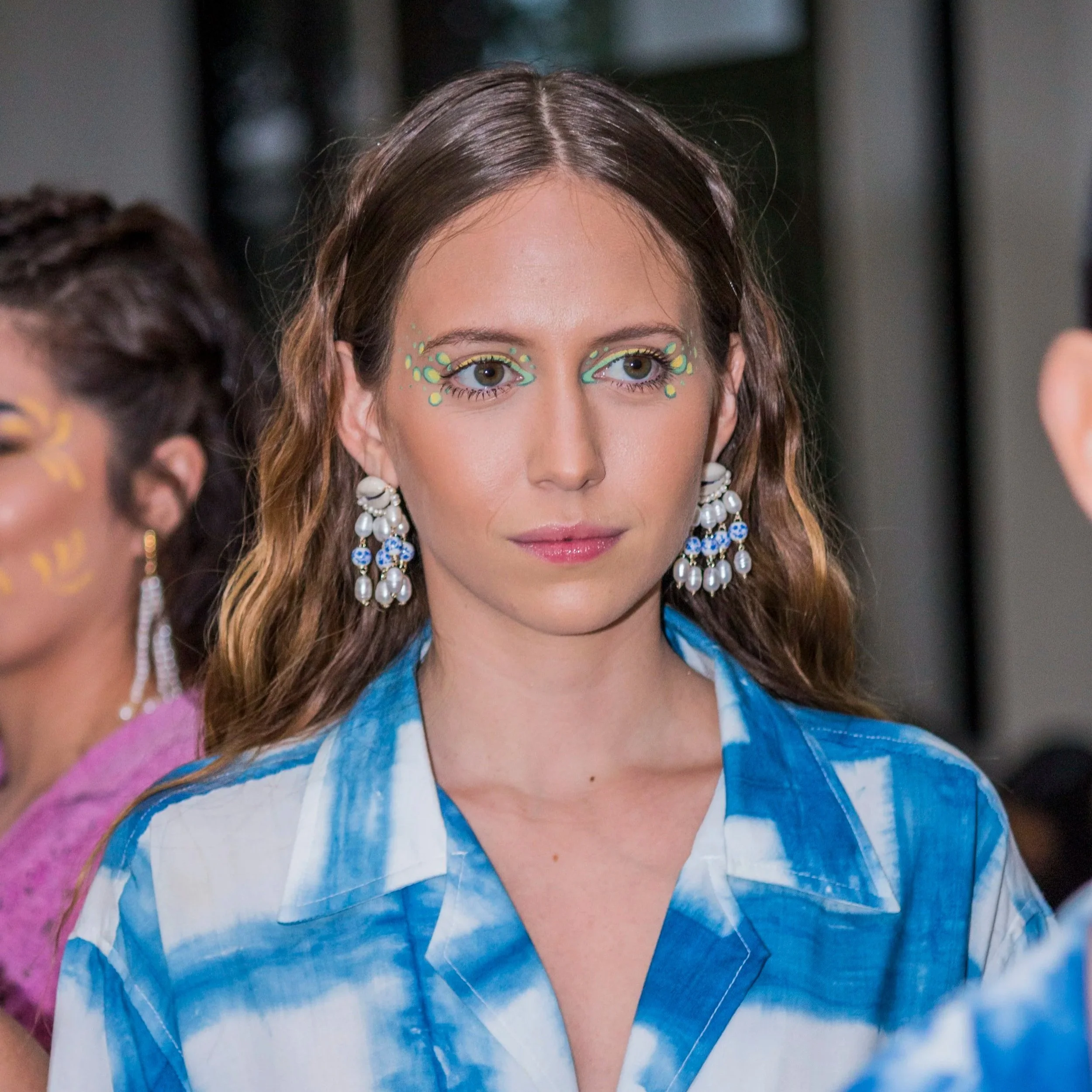 Close-up of a woman with long brown wavy hair and large pearl and blue bead earrings, wearing a blue and white tie-dye shirt, with eye makeup featuring green and yellow designs around her eyes.