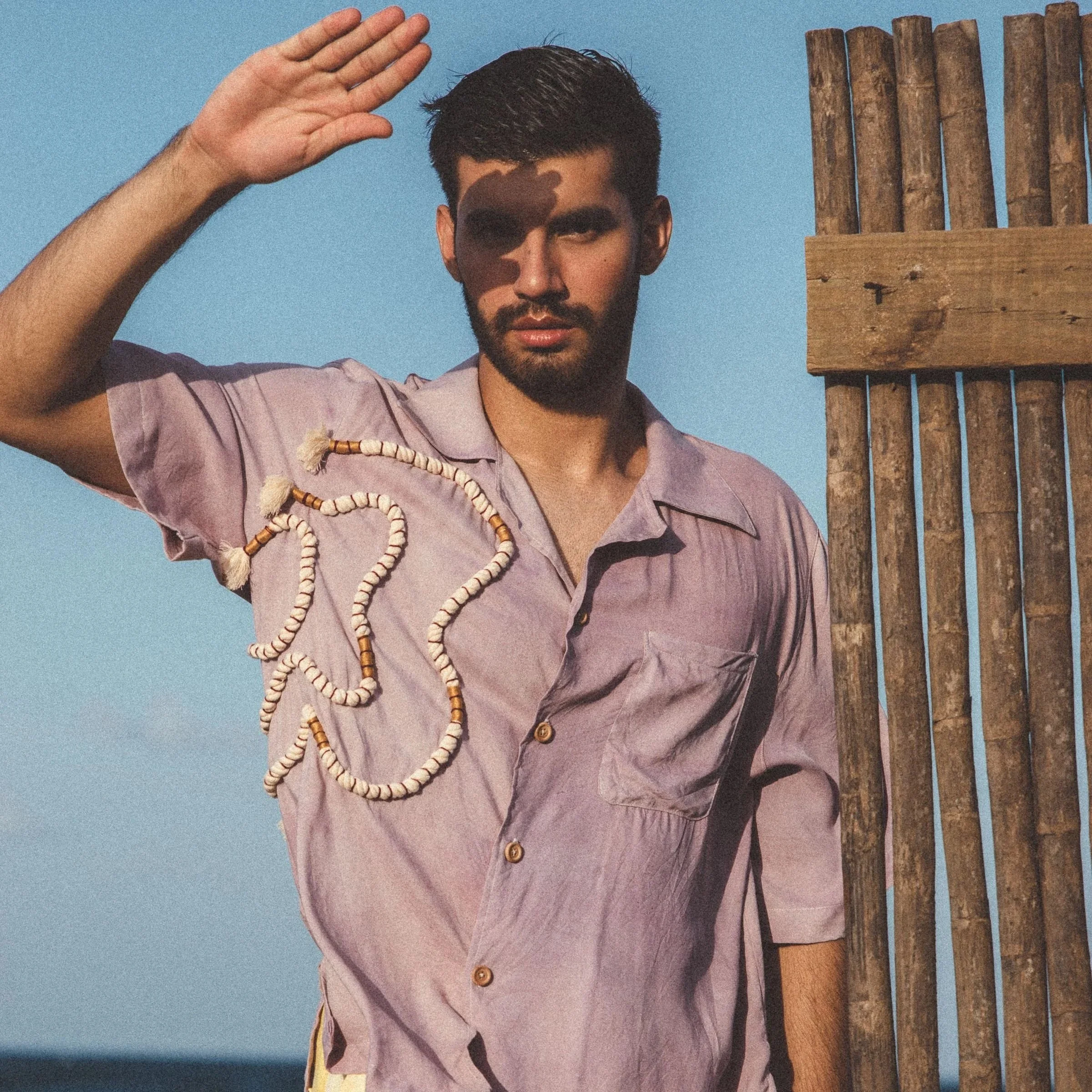 A young man with dark hair and a beard saluting while standing on a beach near a wooden fence, with the ocean and sky in the background. He wears a light purple shirt and yellow shorts.