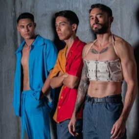 Three young men standing against a plain wall, dressed in casual and eccentric clothing, with serious expressions.