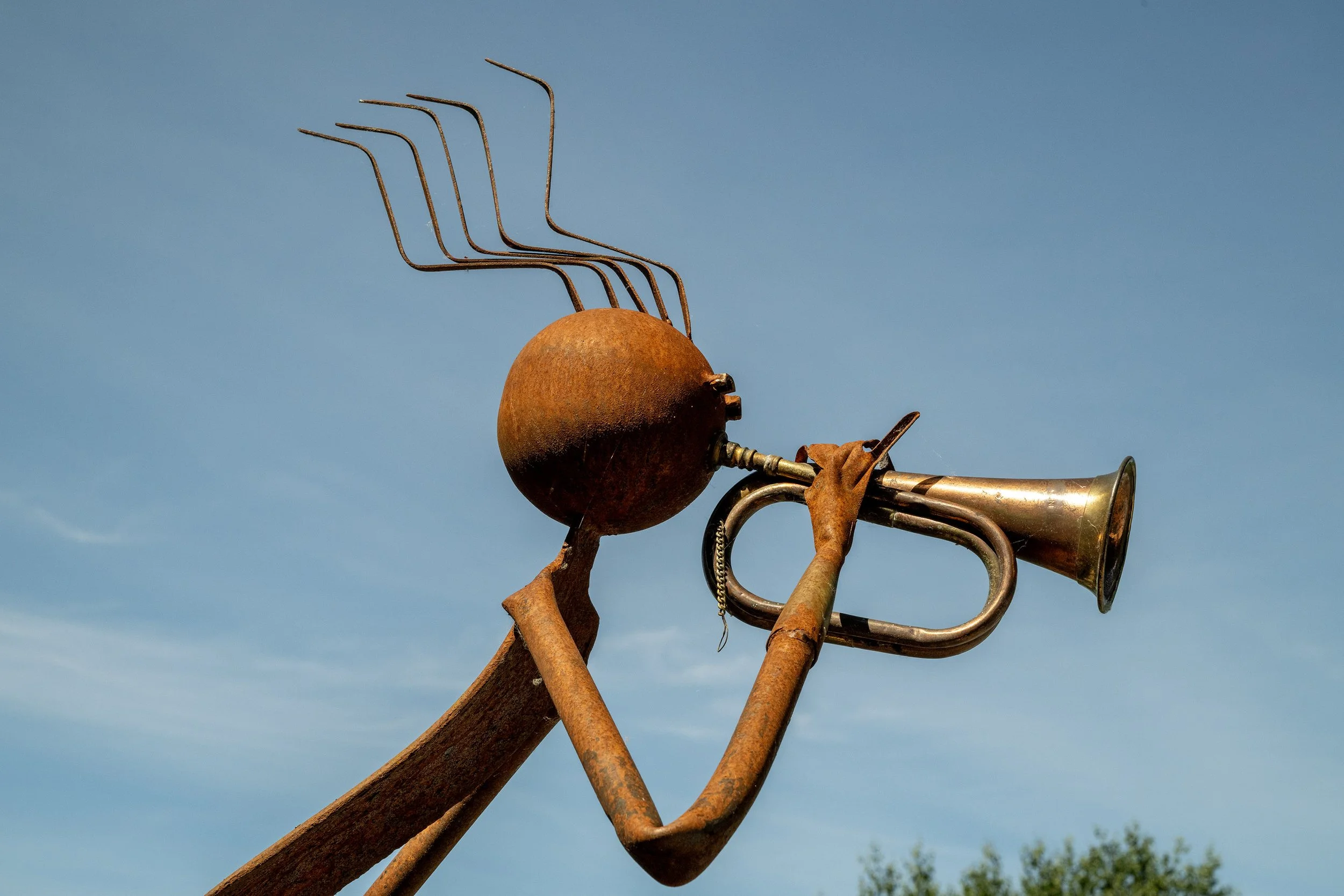 A rusted metal sculpture of a human figure playing a trumpet, with some of the abstract body parts extended upward, set against a clear blue sky.