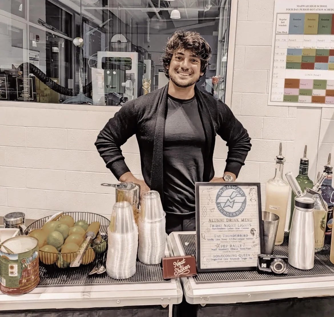 A young man with curly hair and a beard standing behind a drink station at a school event, smiling at the camera. The station includes cups, fruit, bottled drinks, and a framed menu sign.